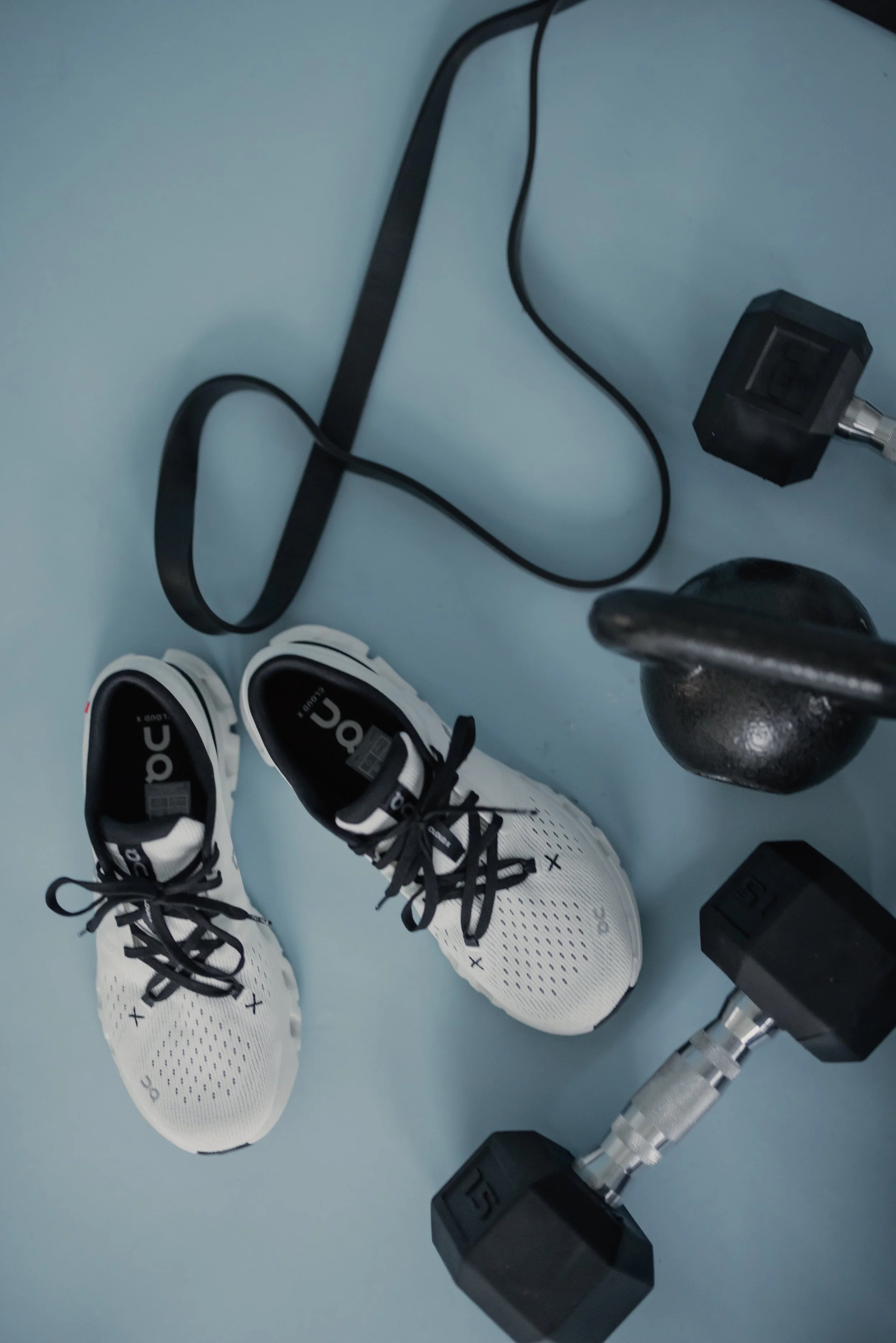 White athletic shoes with black laces, black weights, a kettlebell, and various dumbbells on a light blue gym floor.