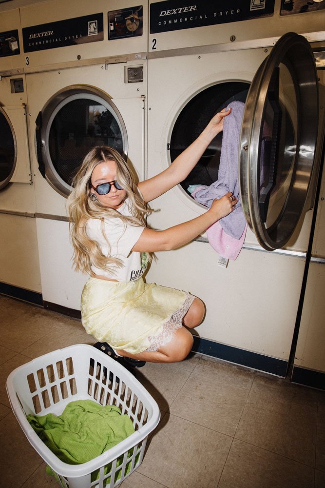 A woman with long blonde hair wearing sunglasses, a white top, and a yellow skirt kneeling in front of a commercial washing machine in a laundromat. She is loading clothes into the machine with a laundry basket filled with green clothes nearby.