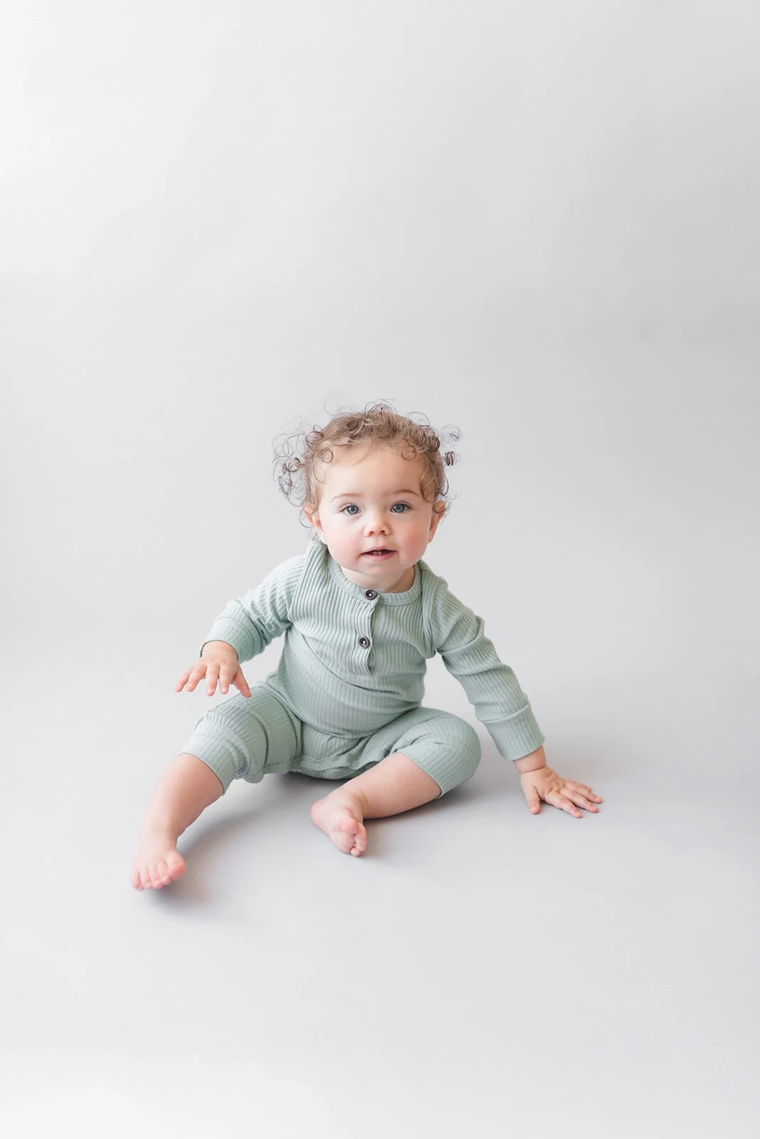 A baby with curly hair sitting on the floor against a plain light gray background, wearing a light green ribbed pajama set with buttons.