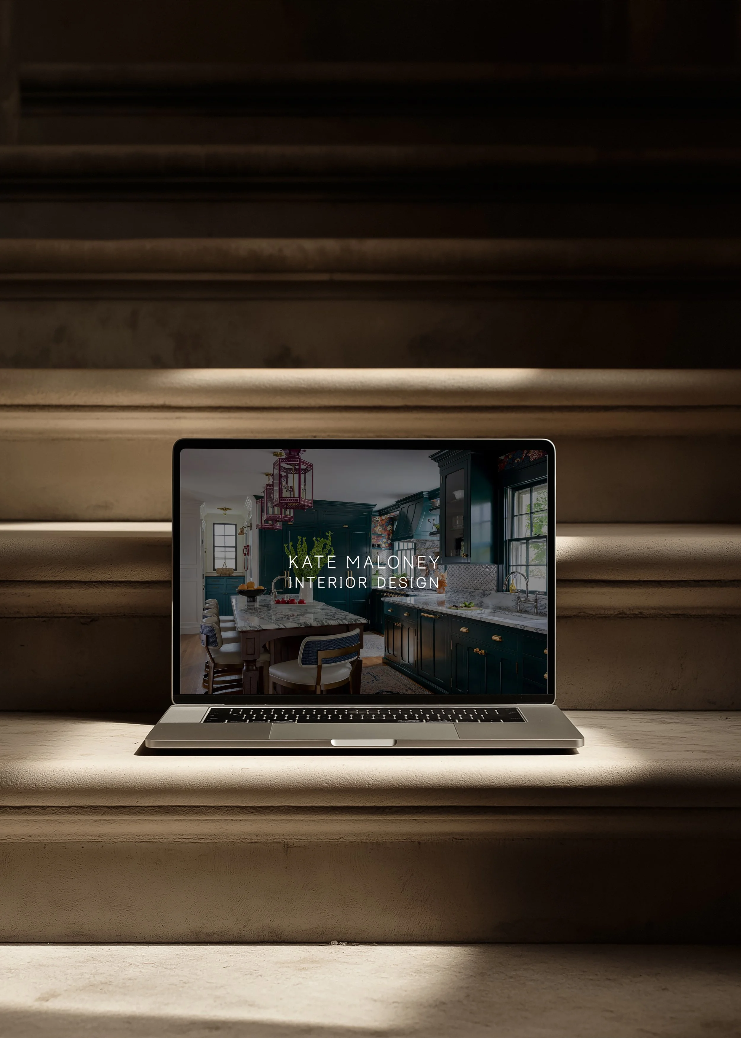 A laptop on a wooden table displaying a kitchen interior design website, with stairs in the background.