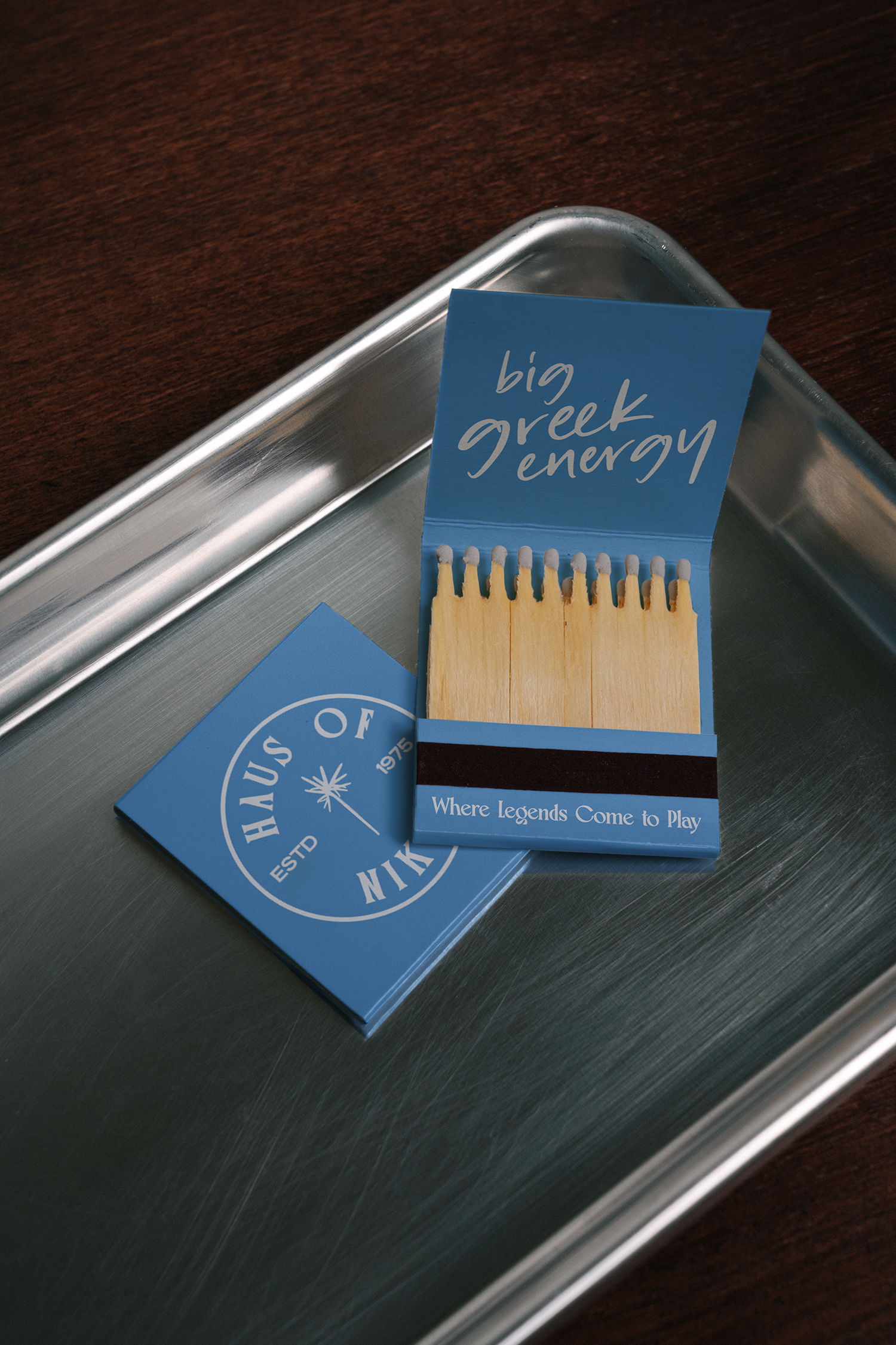 A package of Big Greek Energy matches with a graphic of matches and the text 'big greek energy' on a blue background, placed on a metal tray alongside a blue circular sticker or coaster with white text.