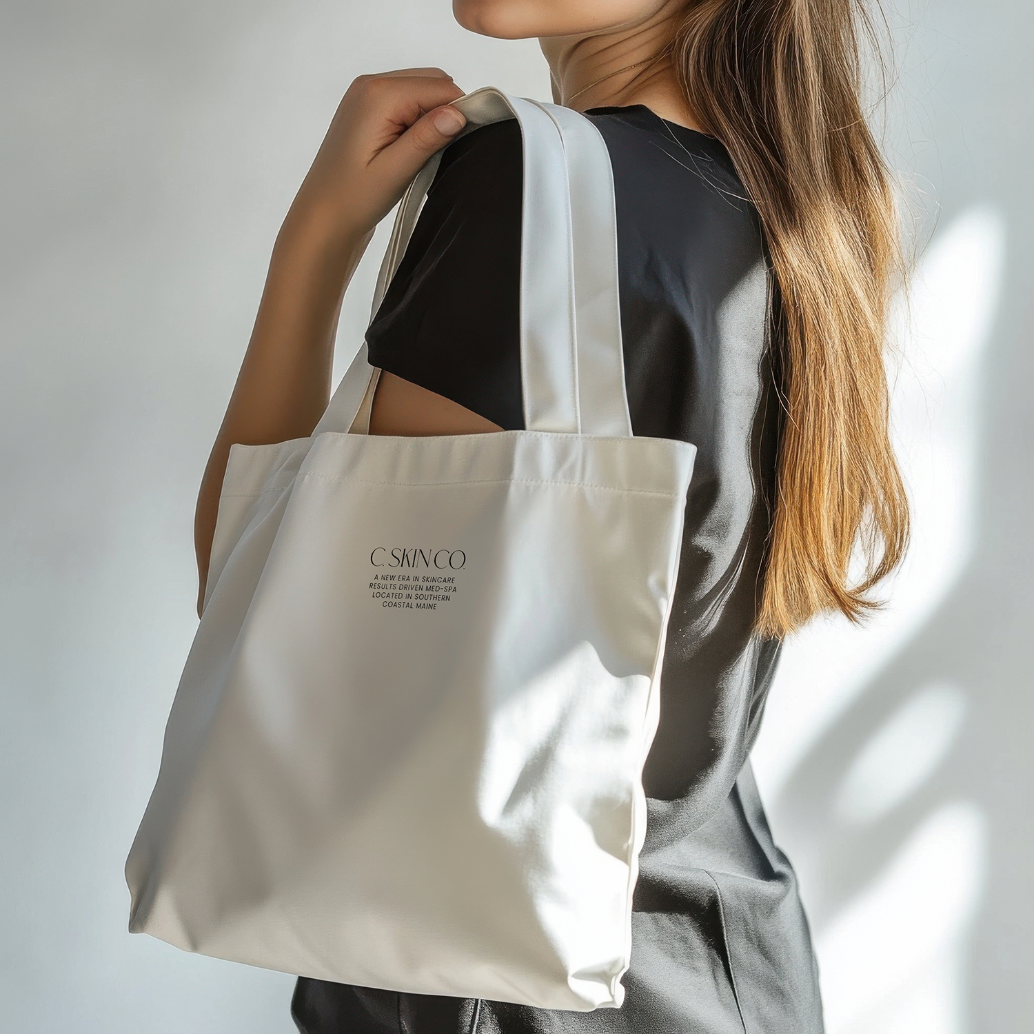 A woman with long, reddish-brown hair is holding a white tote bag over her shoulder. The bag has black text on it that reads 'C. SKINCO' and some additional smaller text. She is wearing a black short-sleeved top and a gray skirt or pants.