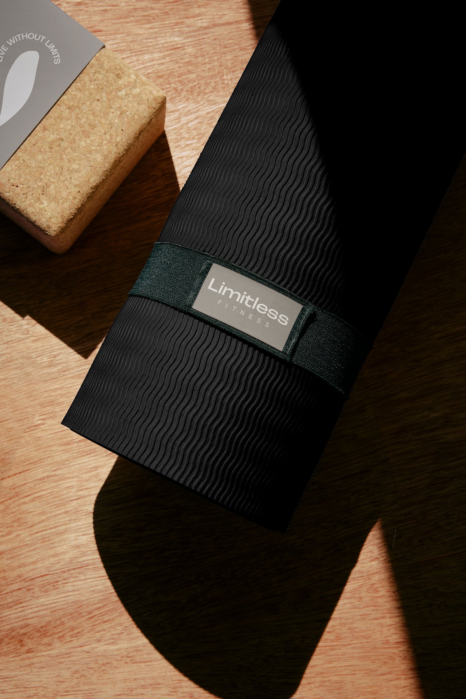 A black fitness mat with the brand name 'Limitless Fitness' on a label, casting a shadow on a wooden floor next to a cork object and a paper with text.