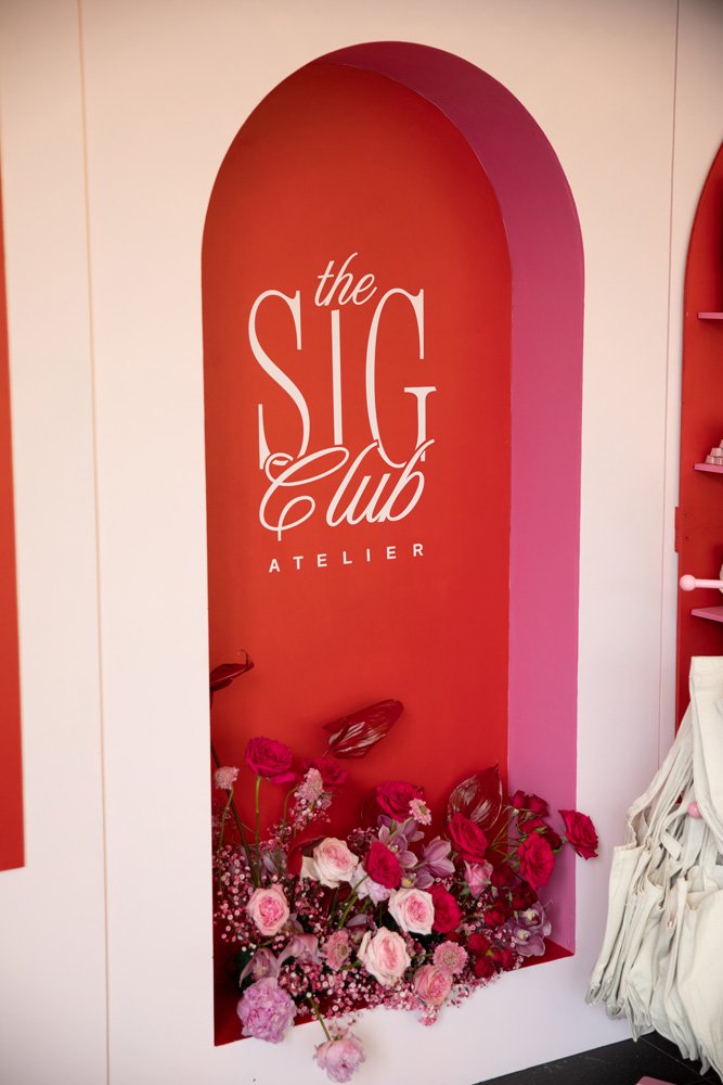 A pink and red wall with an arched cutout displaying the words "the SIG club ATELIER" in white text, with pink and purple flowers arranged at the base.