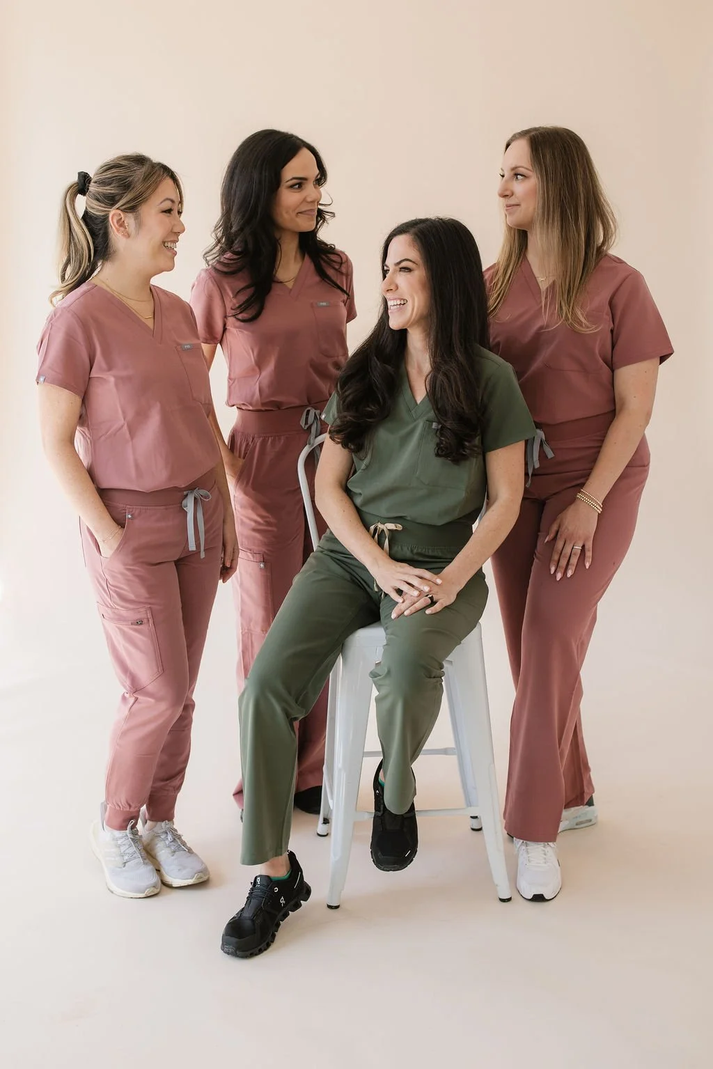 Four women in medical scrubs, one sitting on a stool and the others standing around, smiling and looking at her against a plain beige background.