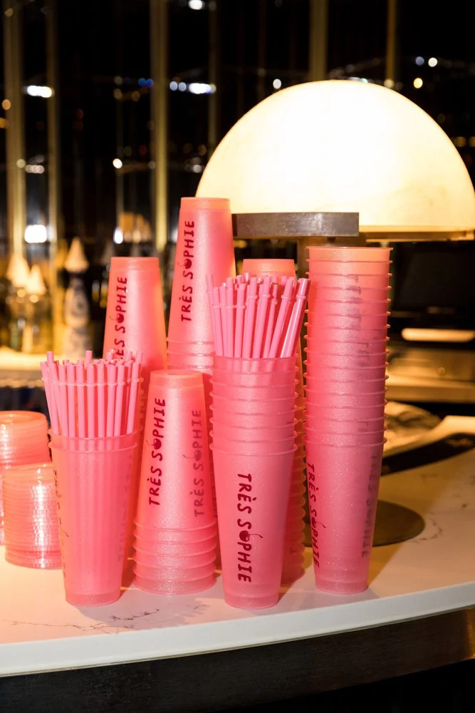 Stacked pink plastic cups with red text that reads "Très Sophie," placed on a white marble countertop illuminated by a warm lamp, with a blurred cityscape in the background.