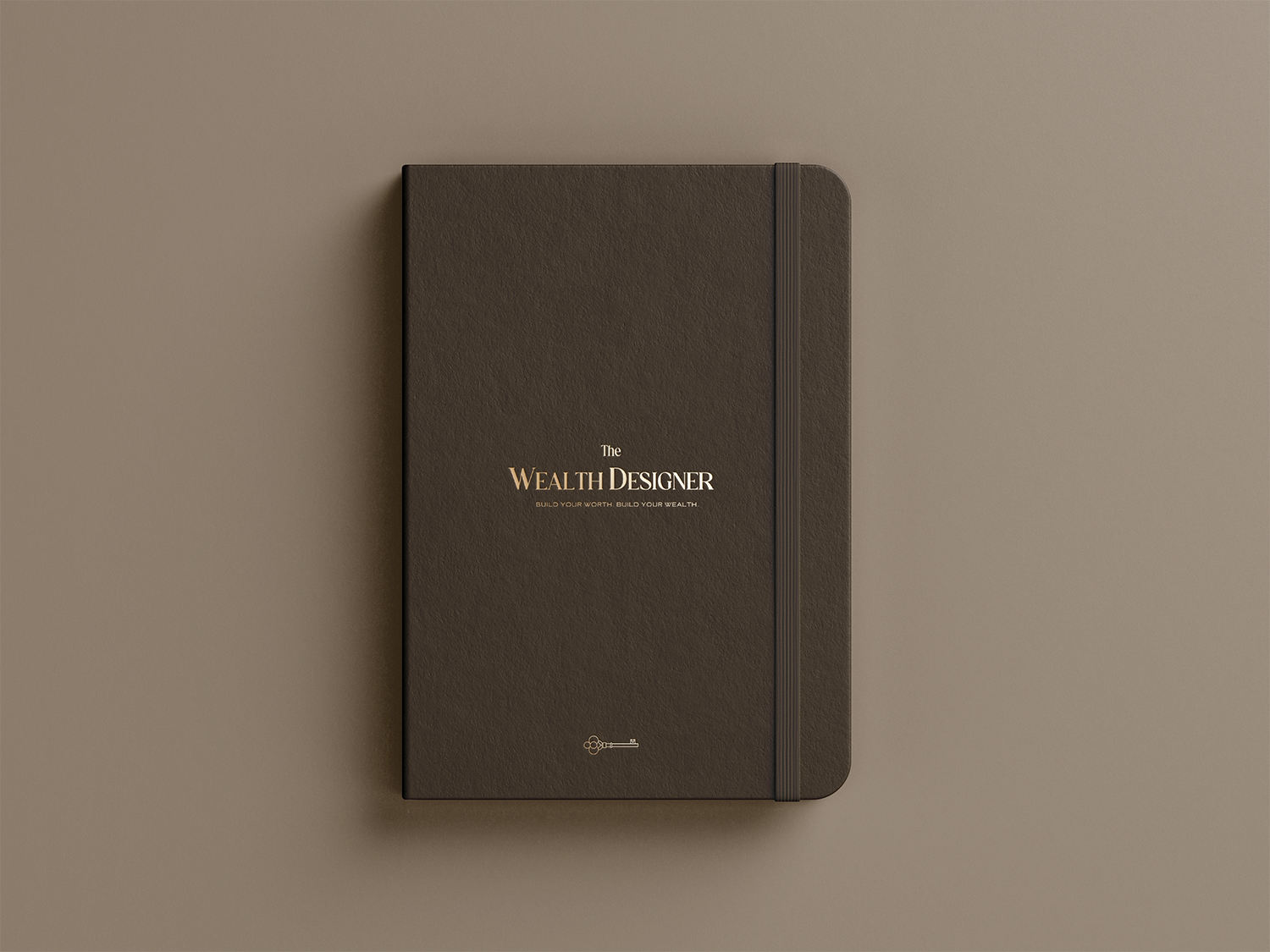 A closed dark brown notebook with gold lettering titled 'The Wealth Designer' and a small key icon on the cover, placed on a beige surface.
