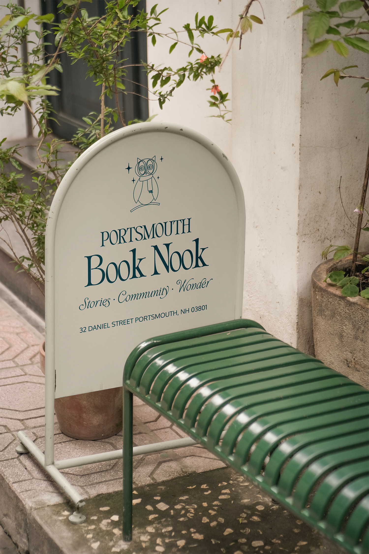 A white sign with blue text reading 'Portsmouth Book Nook' and a simple illustration of an owl, situated next to a green bench outside on a sidewalk, with potted plants and a white wall in the background.