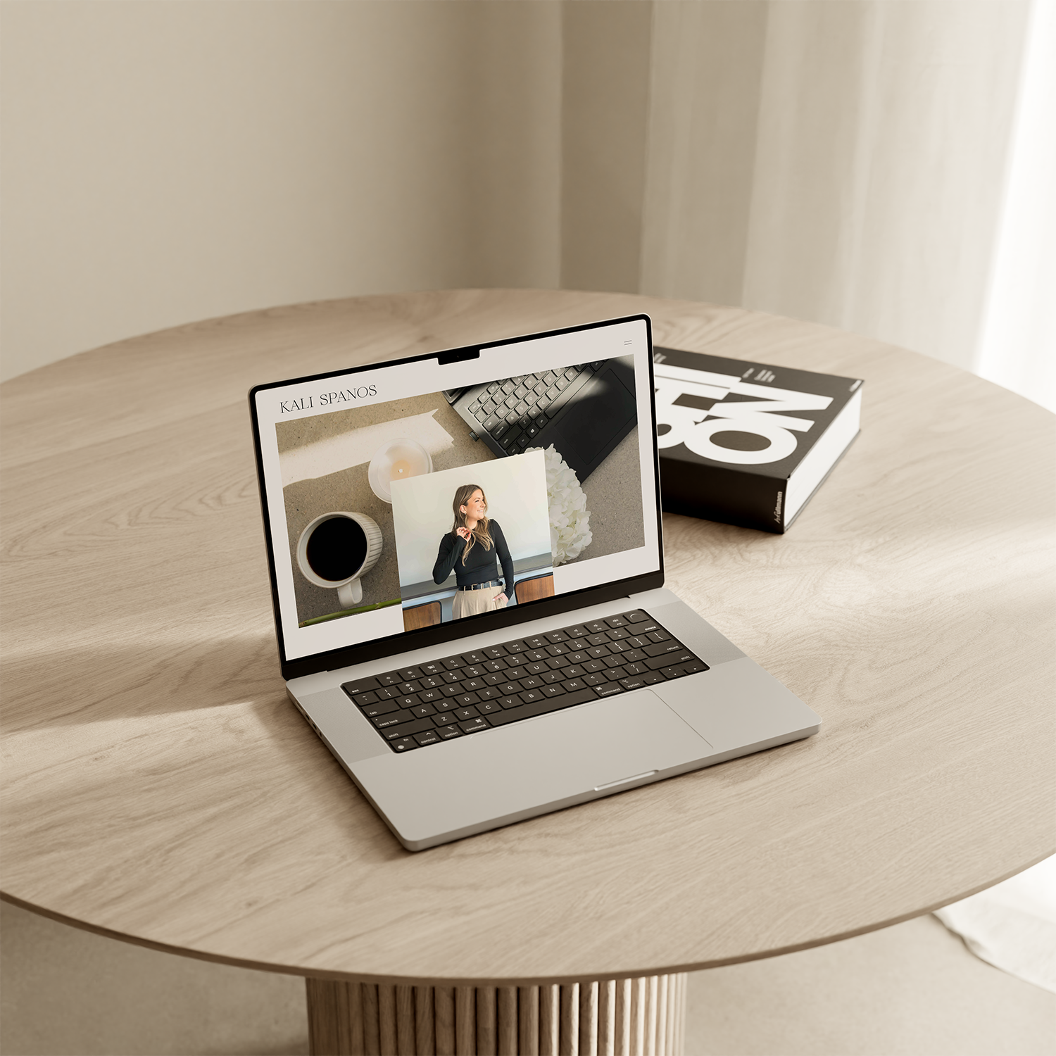 A silver laptop on a light wooden table displaying a webpage with projects, including a photo of a woman smiling, a cup of coffee, a candle, and a laptop keyboard.