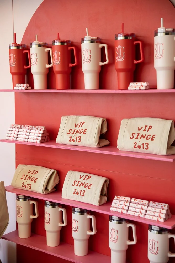Pink bookshelf with large travel mugs, some reading 'the SIG Club' and others read 'VIP SINCE 2013', along with stacks of bar-shaped chocolates.