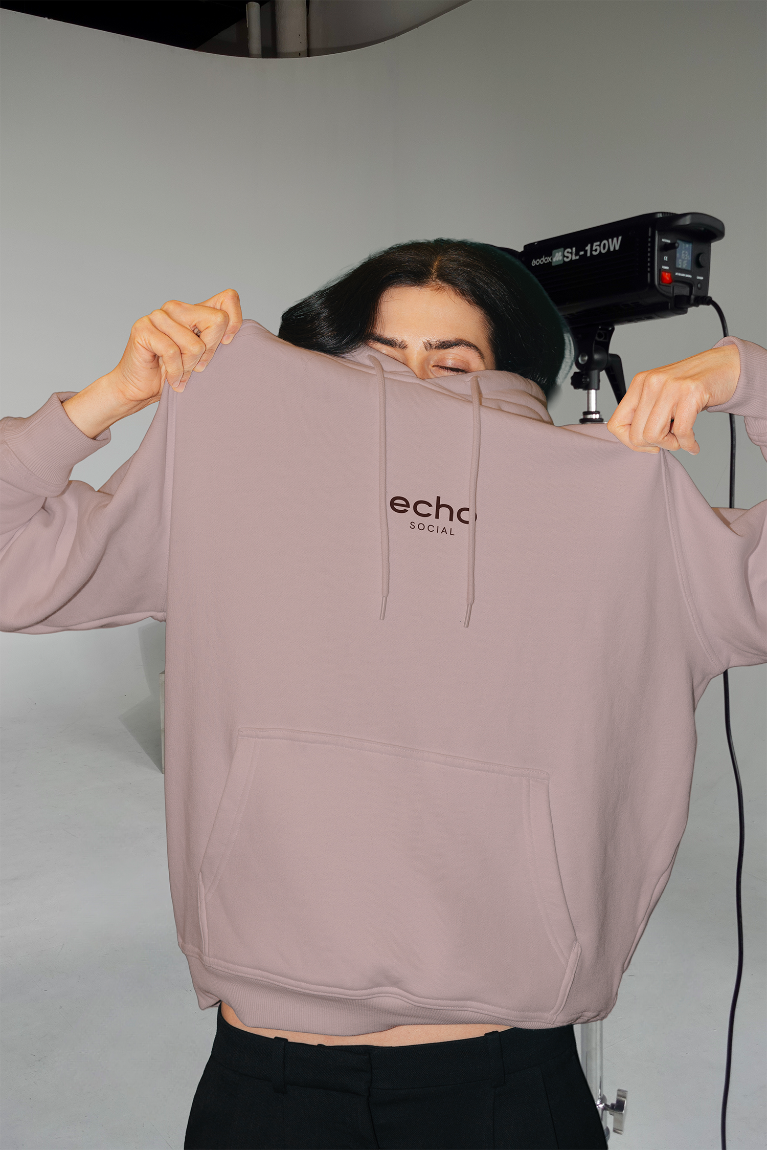 A woman with dark hair, wearing a light pink hoodie that reads 'eche Social,' is pulling the hoodie over her face, revealing only her closed eyes. A photography studio light is positioned behind her, and the background is plain and gray.