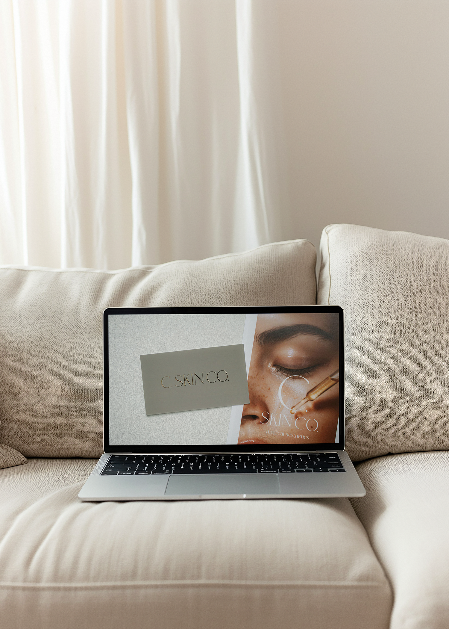 Laptop on beige sofa showing promotional skincare product advertisement, with cream-colored curtains in the background.