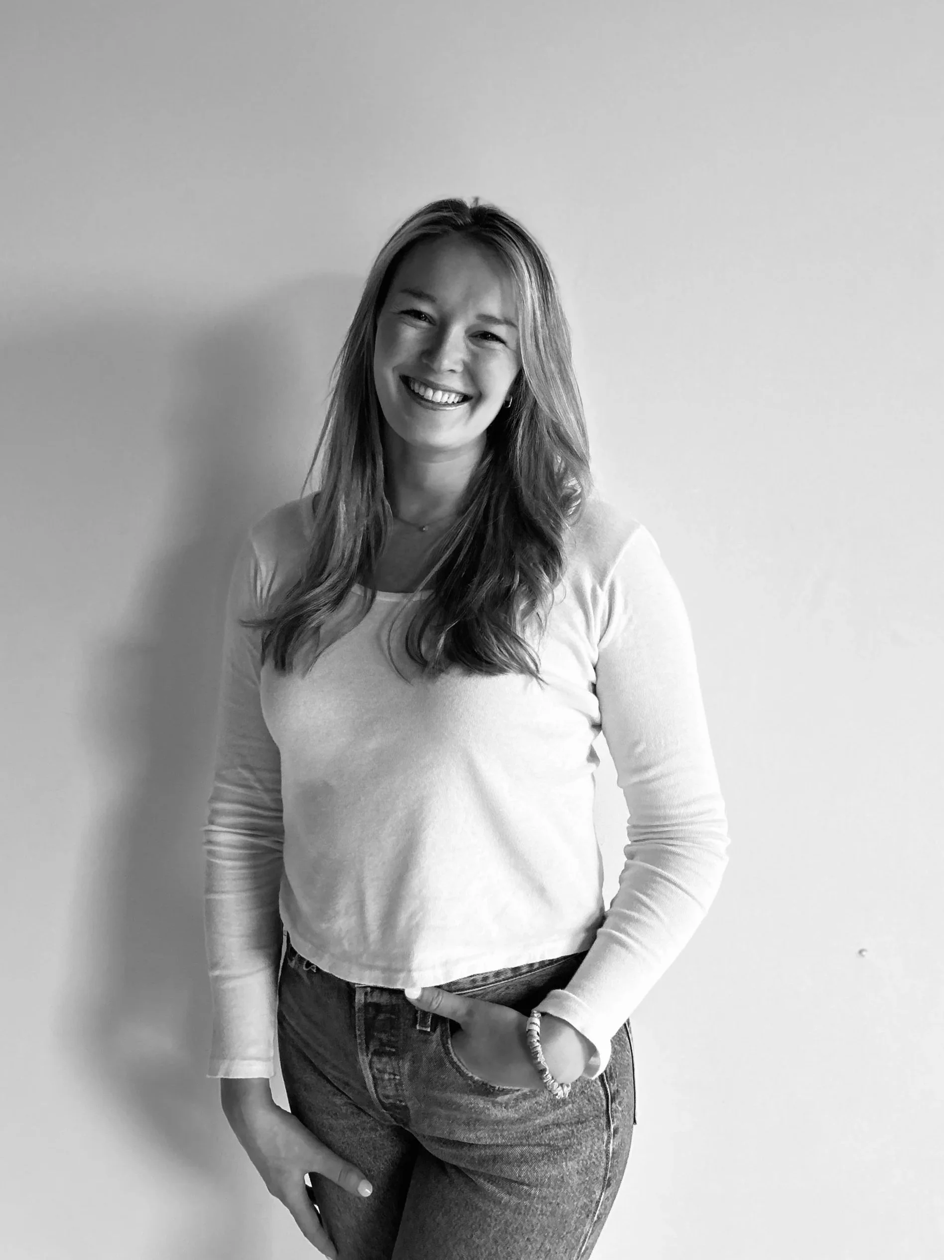 A woman standing against a plain wall, smiling with her head slightly tilted. She has long hair, is wearing a light-colored long-sleeve shirt, and jeans.
