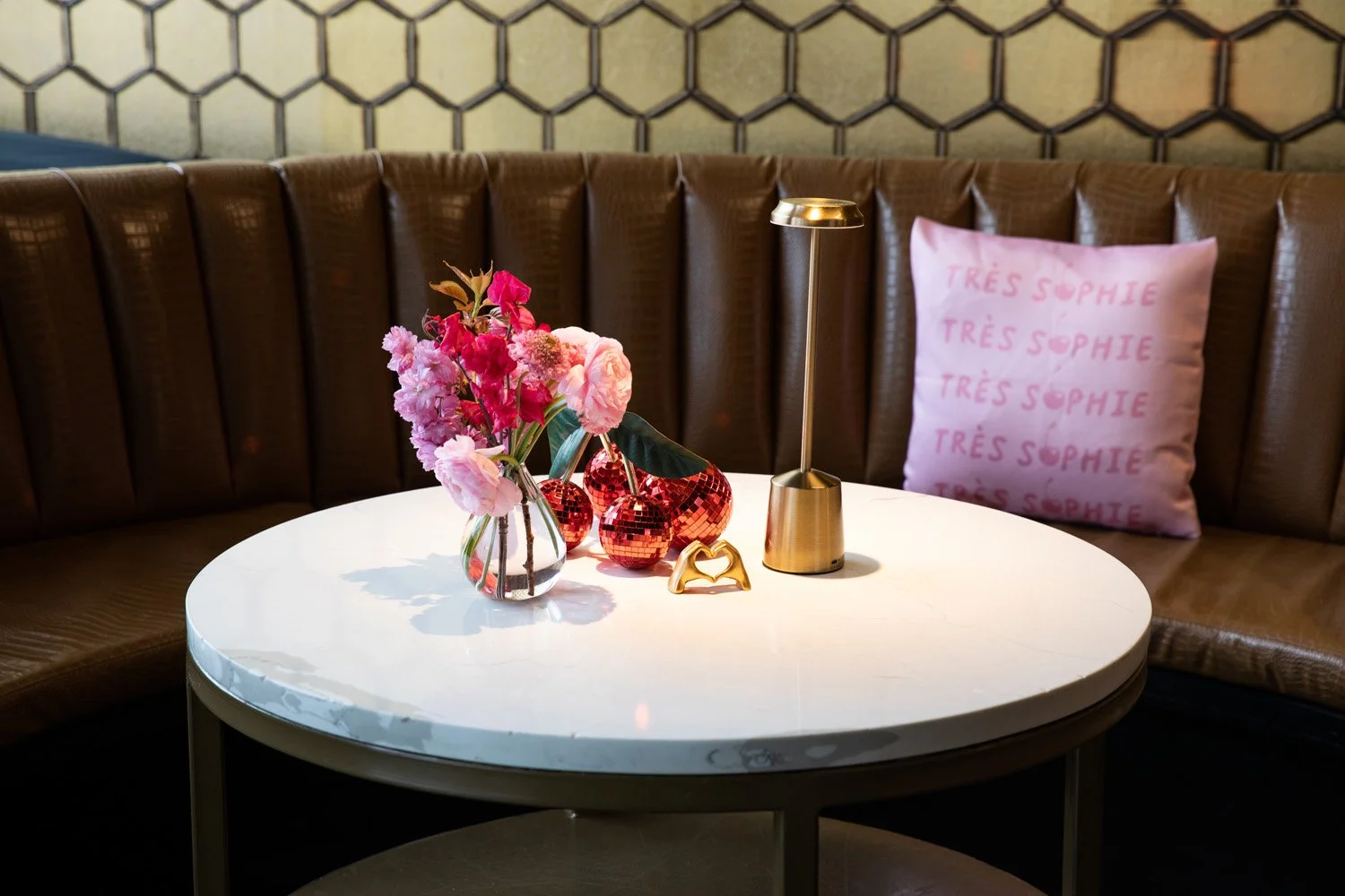 A round white table with a glass vase of pink flowers, three red mirrored vases, a gold desk lamp, and a pink pillow with 'Très Sophie' embroidered repeatedly, in front of a brown leather curved banquette with a hexagonal patterned wall in the background.