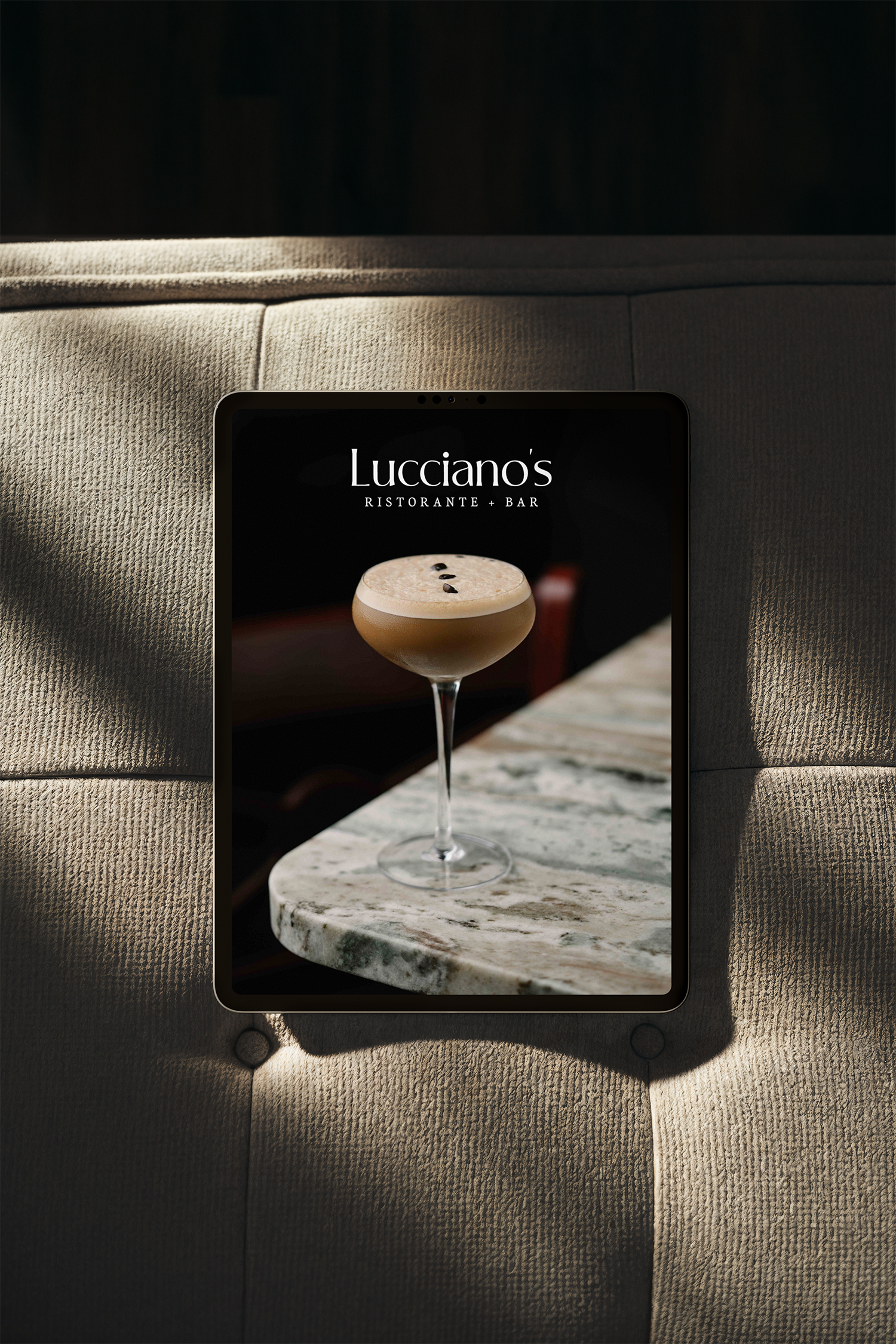 A tablet displaying the logo for Lucciano's Ristorante + Bar, with a cocktail glass on a marble table, and a beige textured sofa in the background.