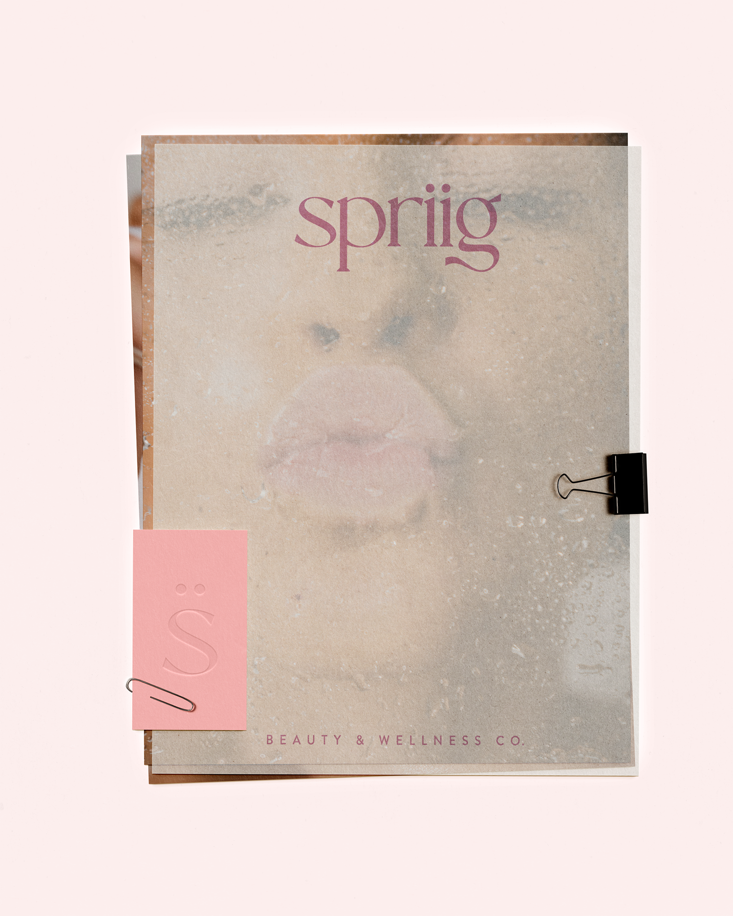 Close-up of an aesthetic beauty and wellness company's branding materials, including a beige textured paper with the word 'spring' in purple and a pink paper clip attached to a pink tab with the letters 'S' in embossed lettering.