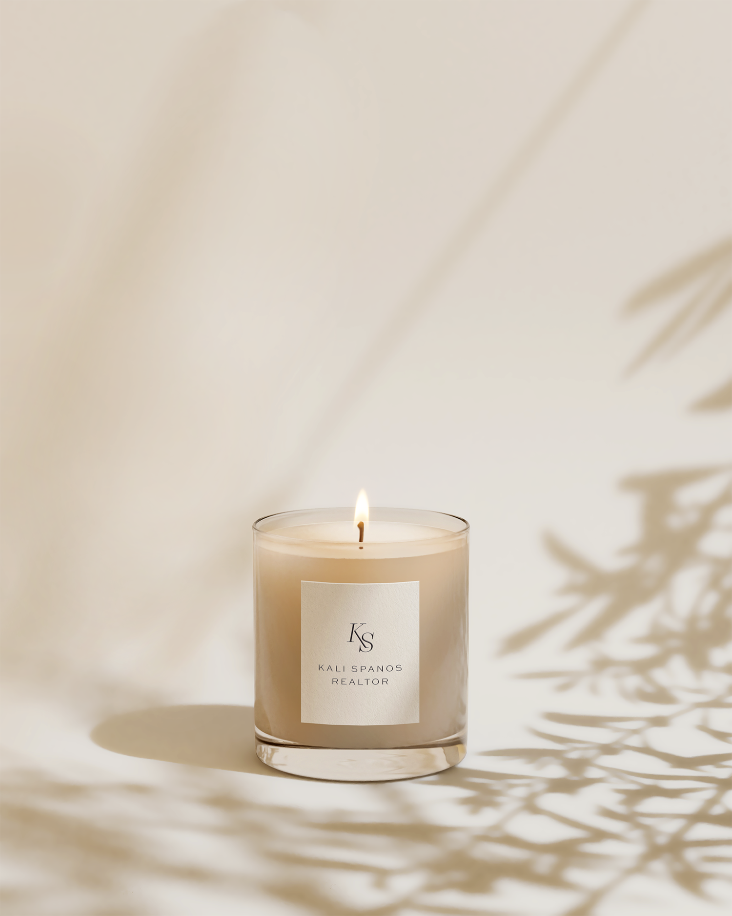 A lit candle in a clear glass container with a white label that reads 'KS Kali Spanos Realtor.' The candle casts shadows of leaves on a white background.