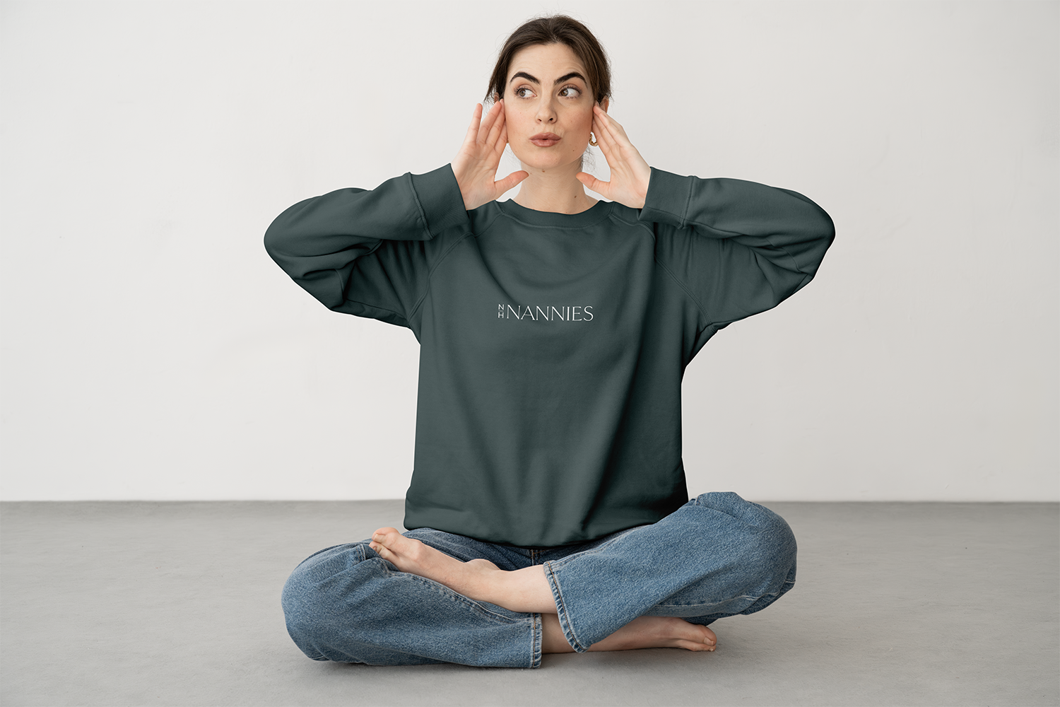 A woman sitting cross-legged on a gray floor, wearing a dark green sweatshirt with 'NANNIES' written on it in white, and blue jeans, touching her face with both hands and looking to the side against a plain white wall.