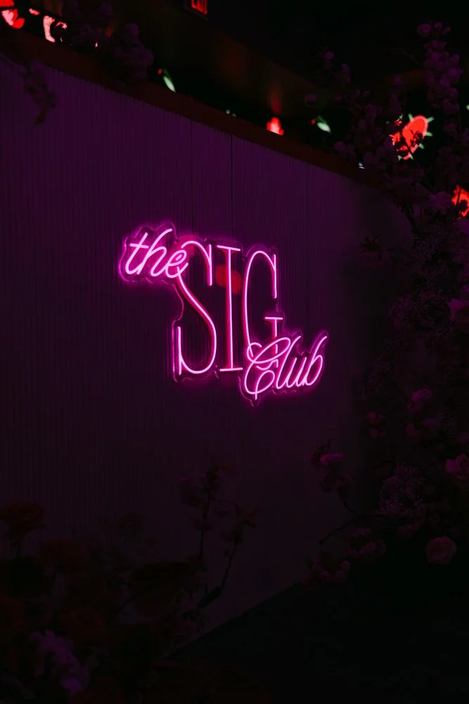 Neon sign reading 'the SIG Club' in pink and purple lights, mounted on a wall with some flowers and dark background.