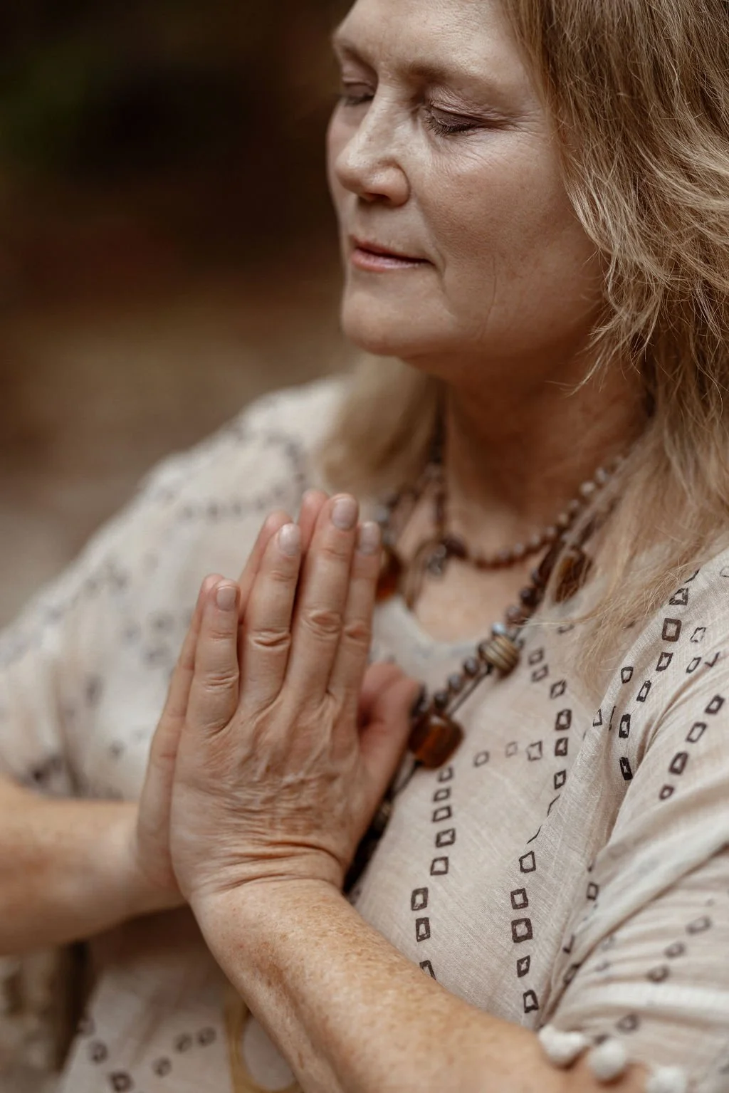 A woman with closed eyes and clasped hands in prayer or meditation, wearing a patterned beige top and layered necklaces, standing outdoors with a peaceful expression.