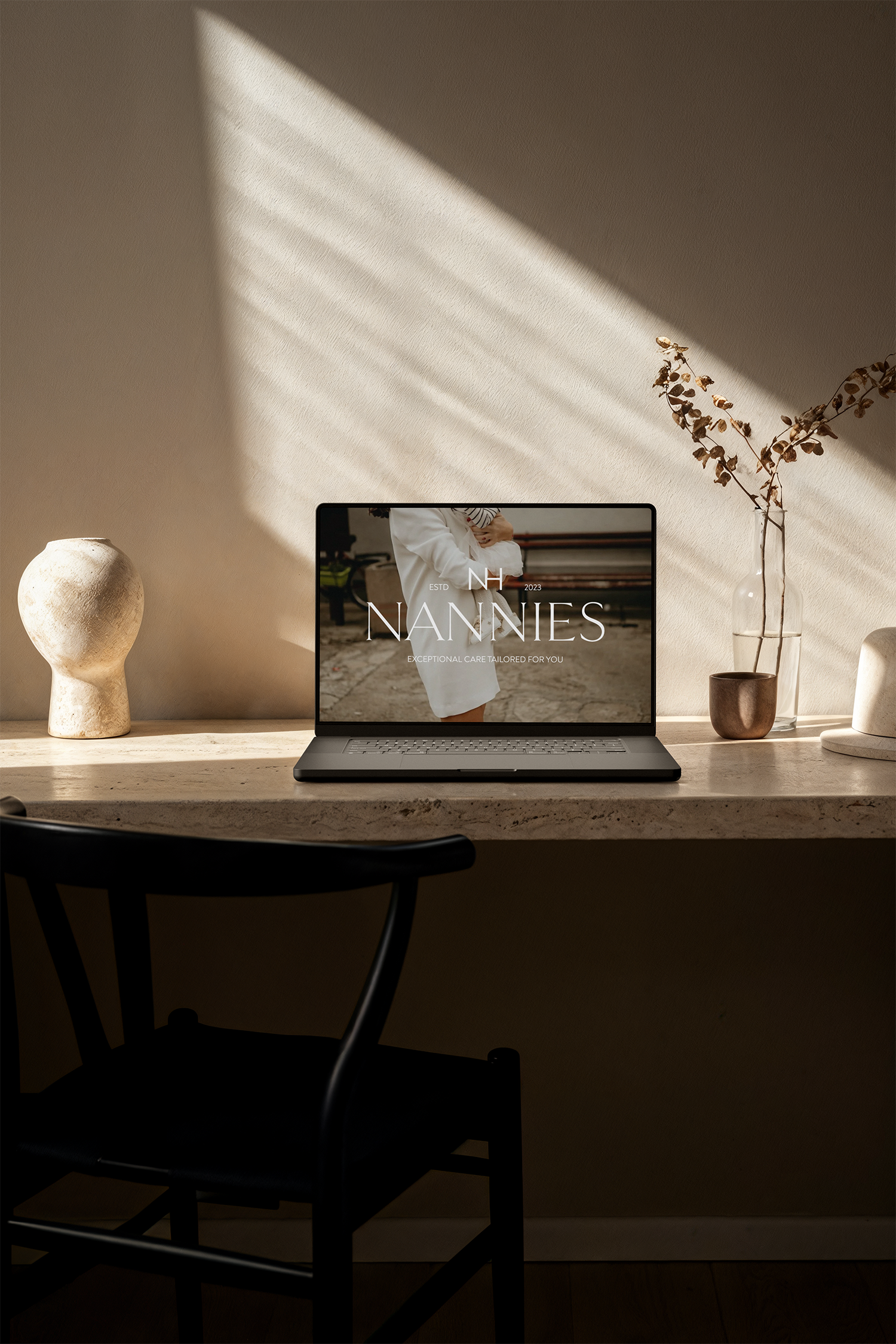 A laptop on a desk displaying a website for Nannies, with sunlight casting diagonal shadows across the beige wall behind it. There are decorative items on the desk, including a textured white vase, a clear glass bottle with dried flowers, and a small