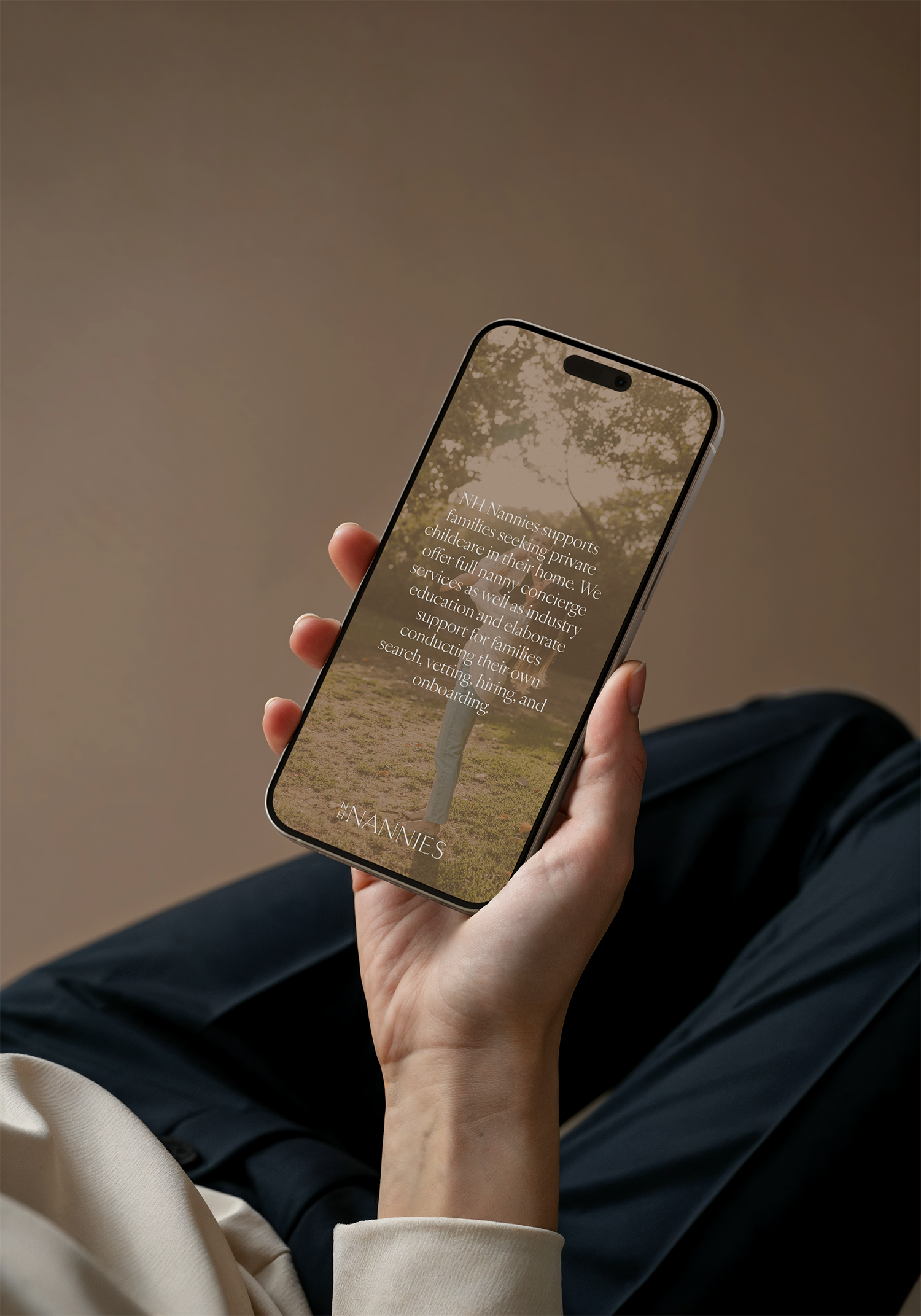 Close-up of a hand holding a smartphone displaying the NHannies supports families seeking private childcare services at home, with a background of trees and natural scenery.