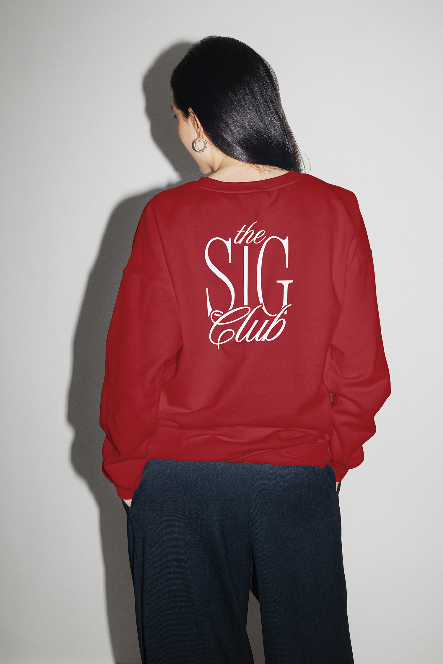 Back view of a woman wearing a red sweatshirt with 'The SIG Club' written on the back, and black pants, standing against a plain light-colored wall.