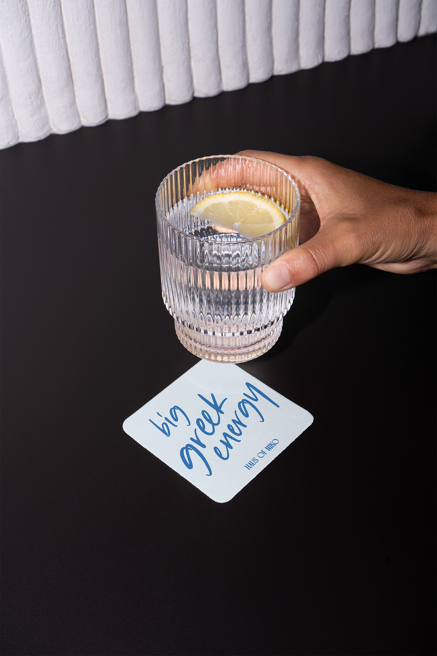 A hand holding a glass of water with a lemon wedge on a dark surface. There is a white piece of paper with blue cursive writing that says 'big love forever' and a logo 'Haus of Niko' at the bottom.
