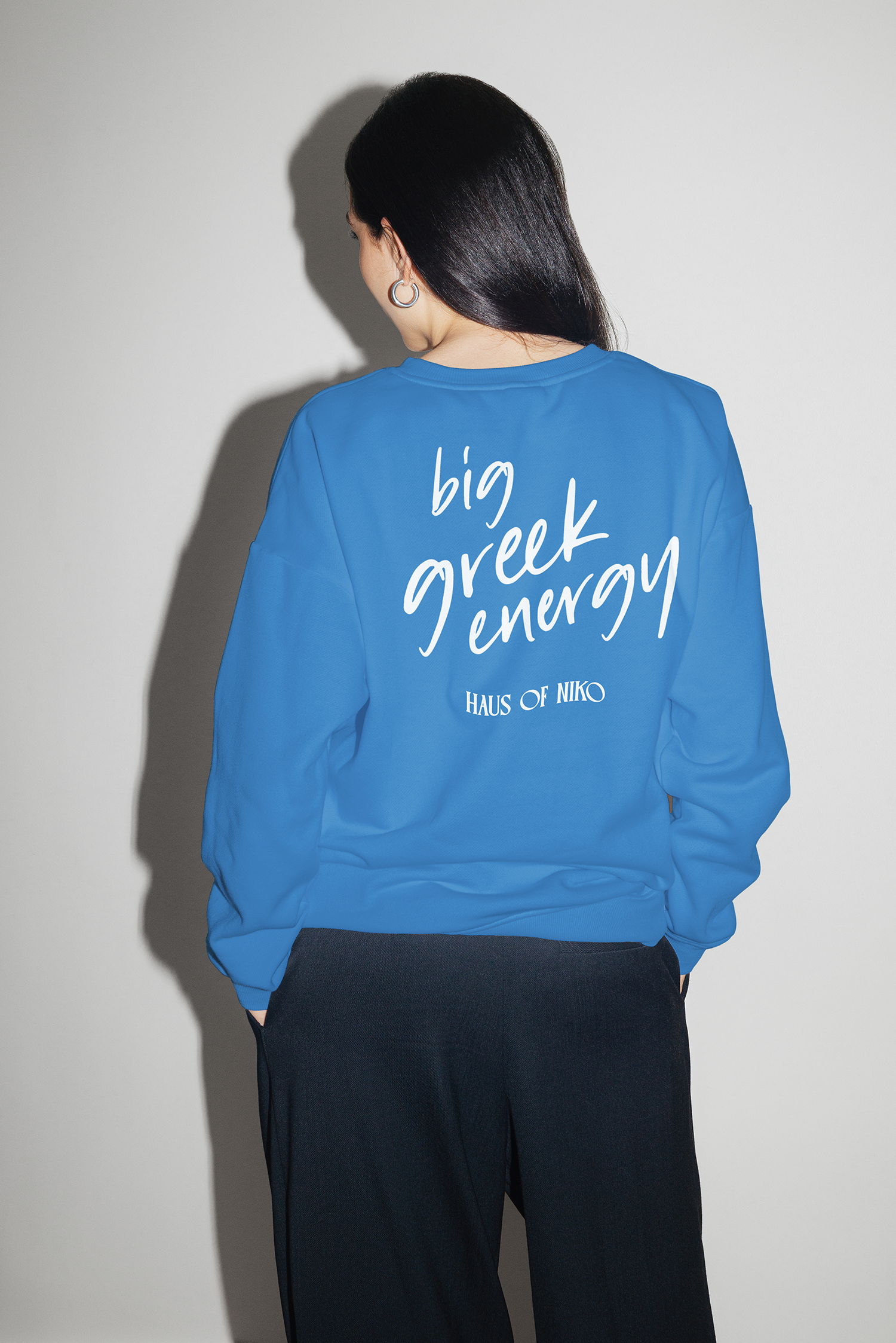 Back view of a woman with long black hair wearing a blue sweatshirt with white text that says 'big greek energy' and smaller text that says 'HAUS OF NIKO', standing against a plain light gray background.