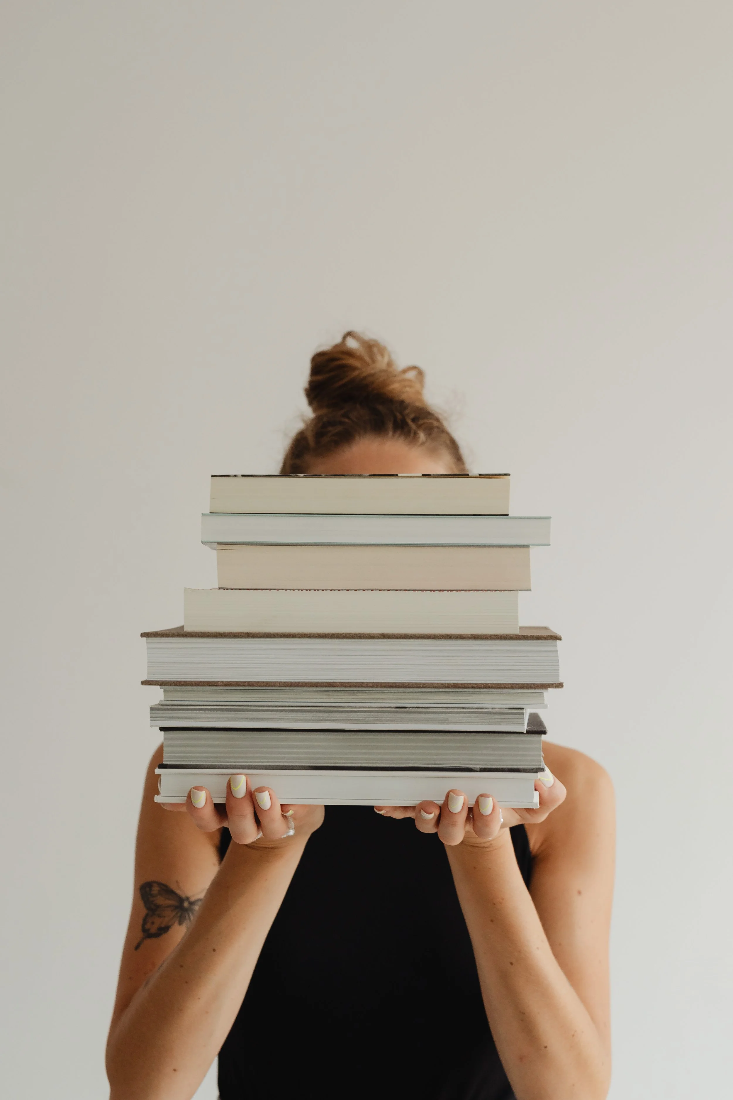 A person with red hair in a bun, wearing a black top, is partially hidden behind a stack of books. They are holding the books in front of their face, and the books obscure their facial features.
