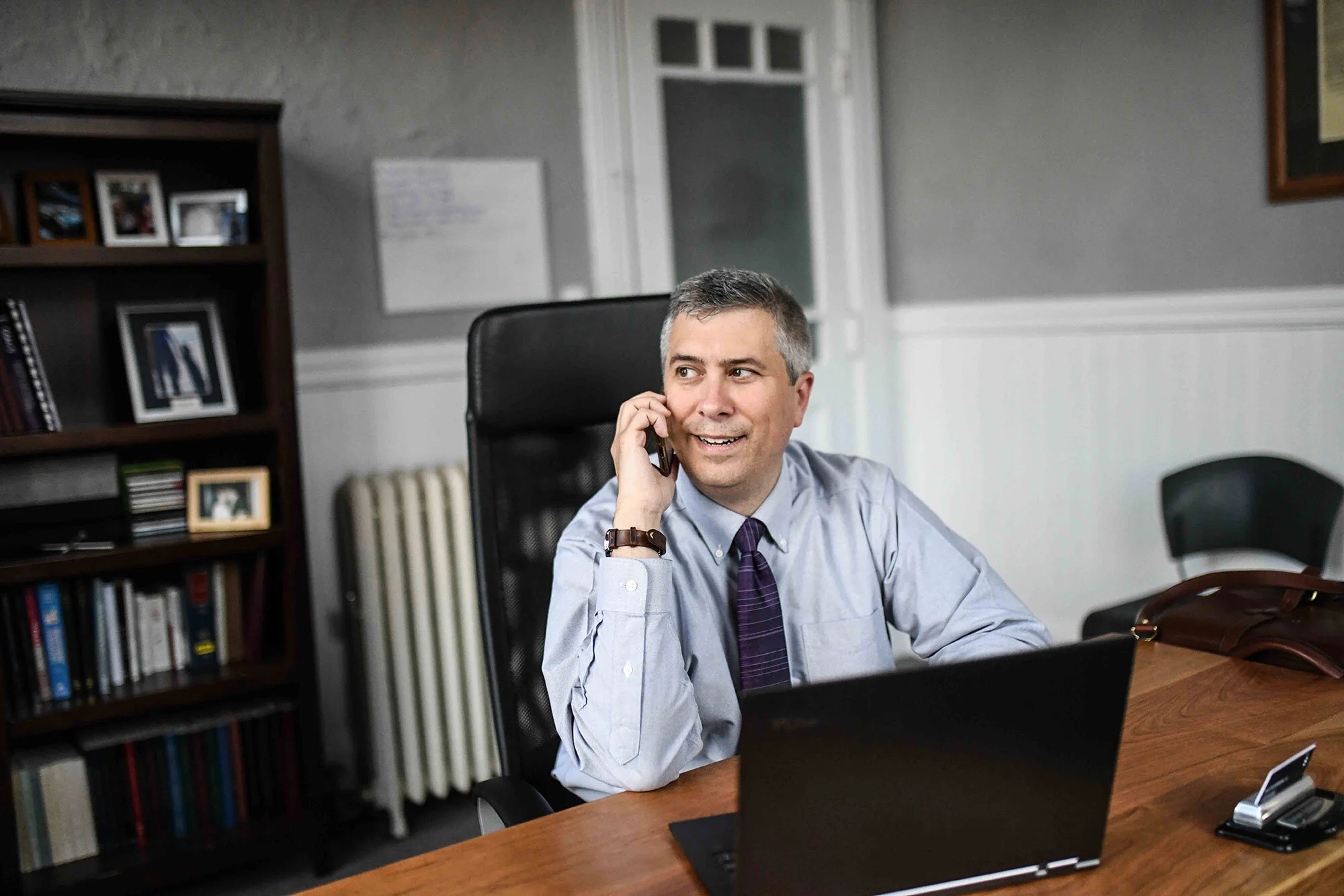Man in office sitting at a desk talking on a phone with a laptop in front of him. Bookshelf and whiteboard in the background.