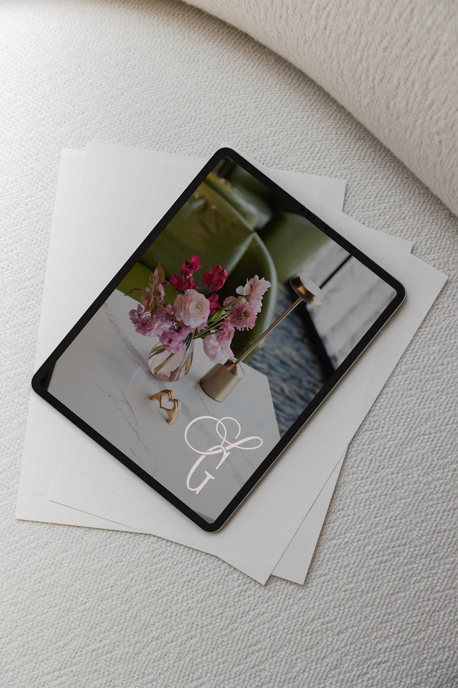 Close-up of a tablet displaying a floral arrangement and jewelry, resting on printed papers on a textured white surface.