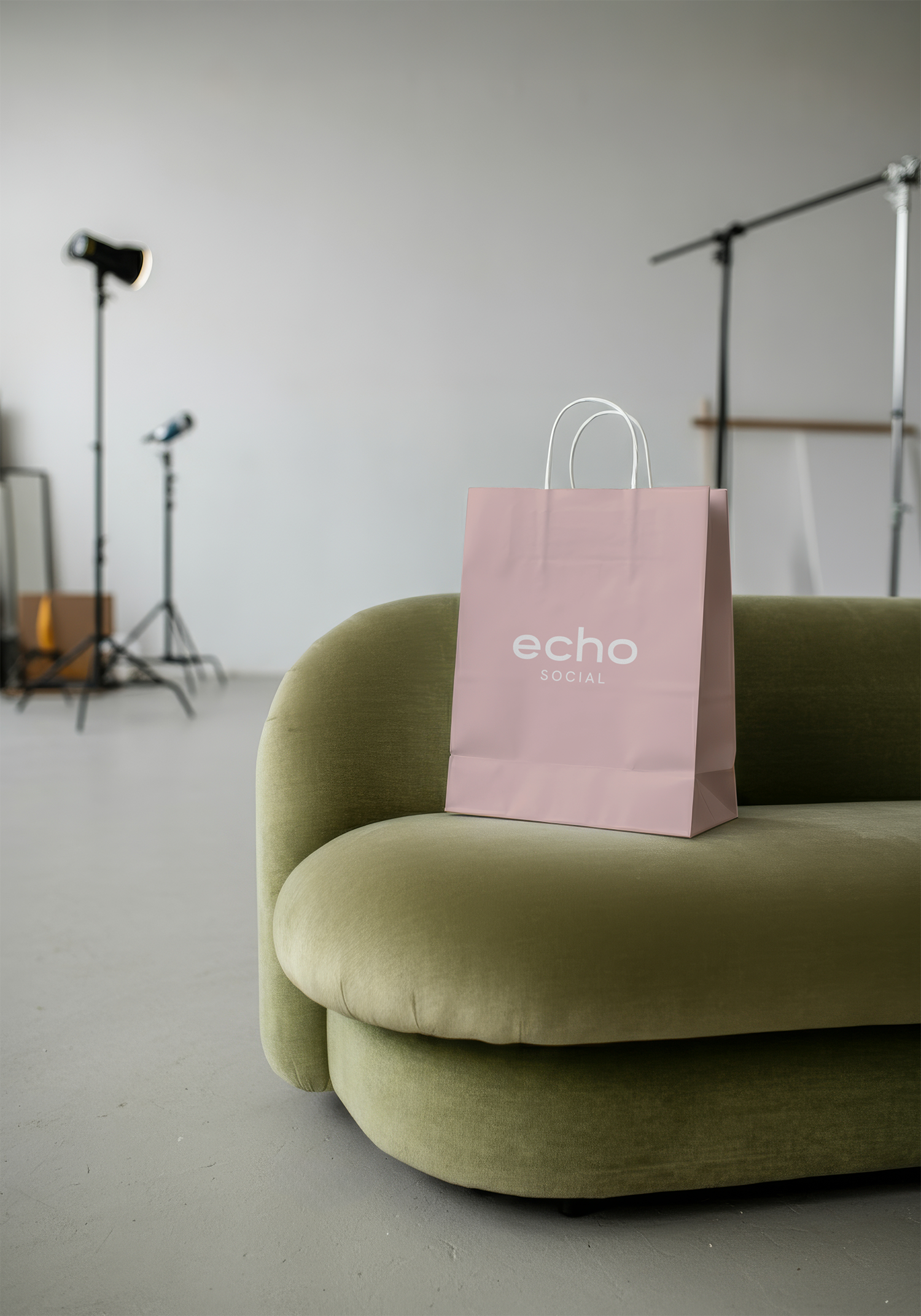 A pink shopping bag with white handles and the words "echo SOCIAL" printed on it, sitting on a green upholstered armchair in a photography studio with lighting equipment and a plain white backdrop in the background.