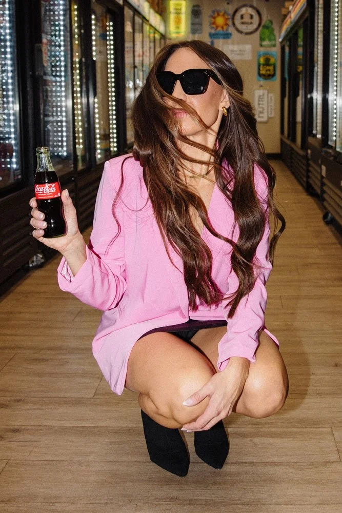 A woman with long brown hair wearing large black sunglasses, a pink blazer, black shorts, and black boots, is crouching in a convenience store aisle, holding a small glass Coca-Cola bottle in her right hand. She is looking at the camera with her hair