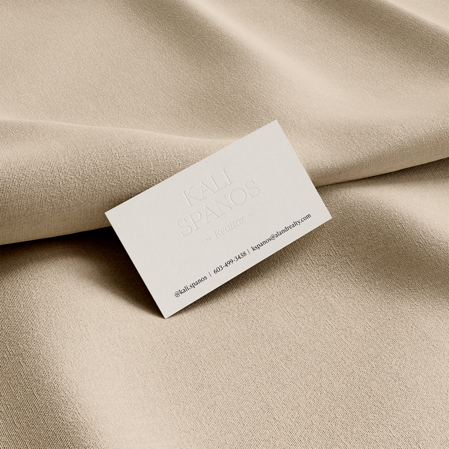 A white business card resting on a beige textured fabric surface. The card has embossed text with the name 'Kali Spanos,' the title 'Realtor,' and an email address, phone number, and social media handle.