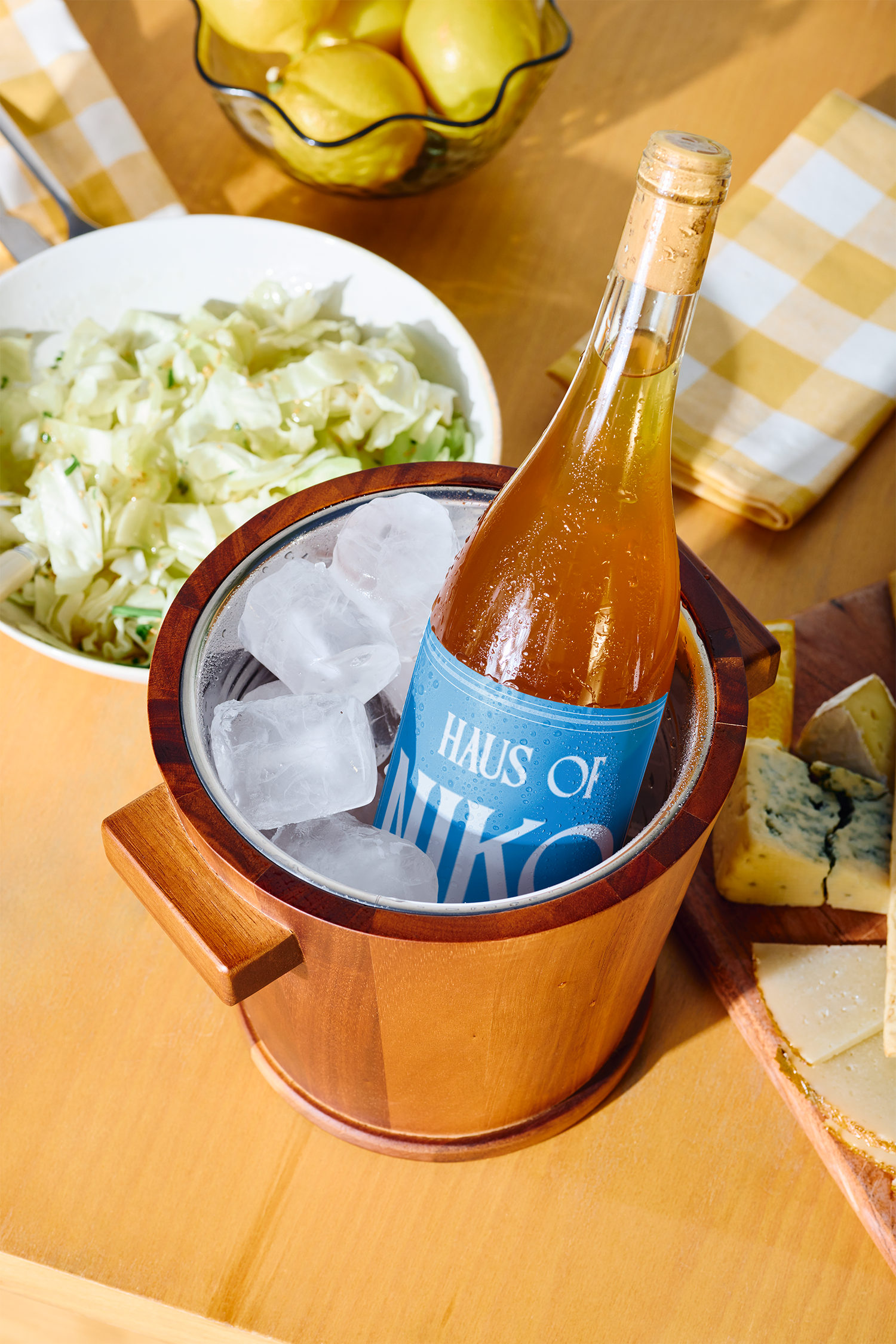 A bottle of sparkling wine in an ice bucket, surrounded by a bowl of lemons, a bowl of coleslaw, and cheese on a wooden table with checkered napkin.