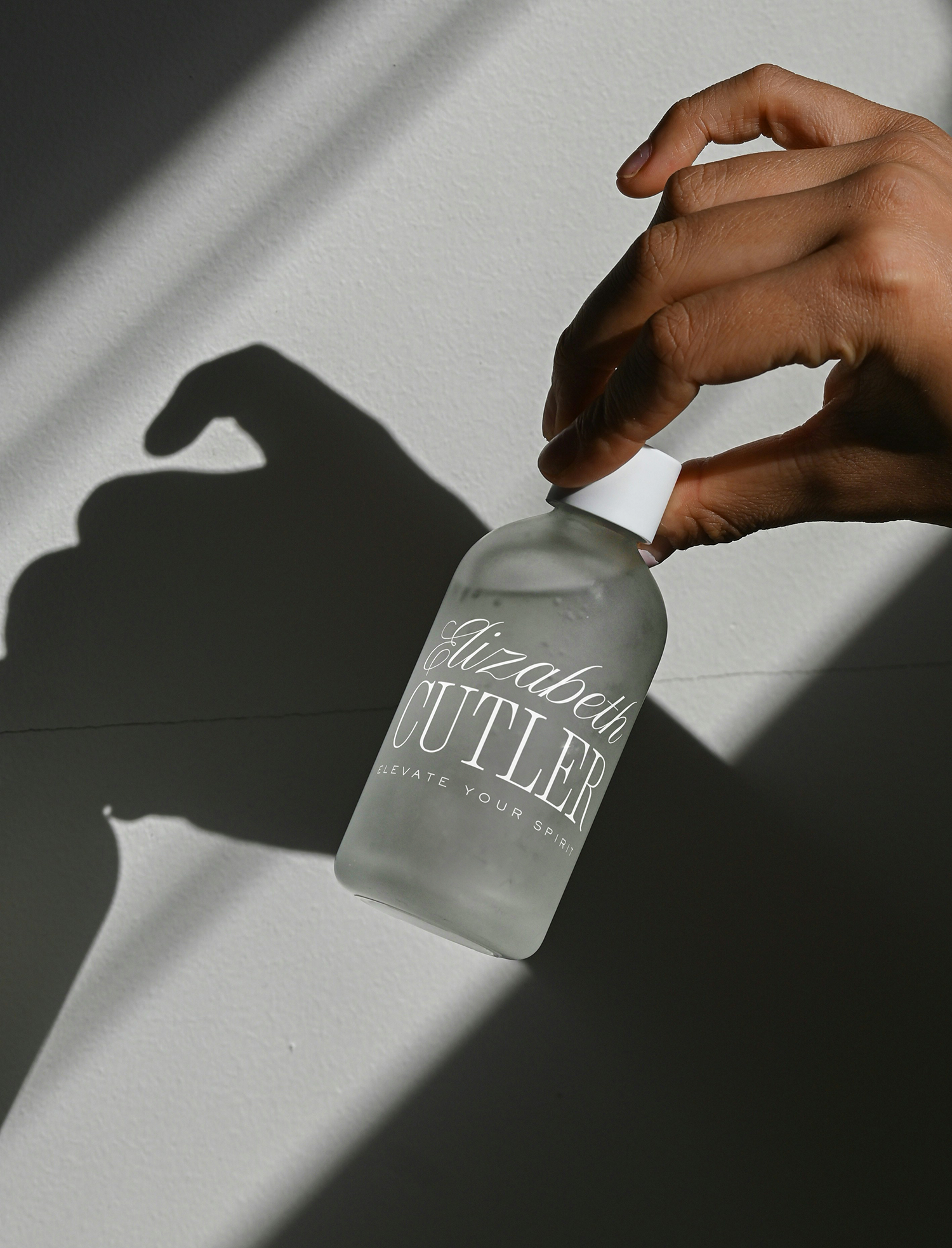 A hand pressing a white pump on a bottle of Elizabeth Cutter skincare product, casting a shadow on a light-colored surface.