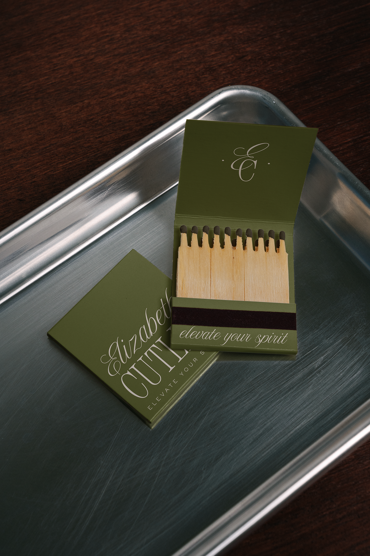 Set of matchbooks with green covers and black stripes, placed on a silver tray on a wooden surface. The covers have elegant script text 'elevate your spirit' and a monogram 'E C'.