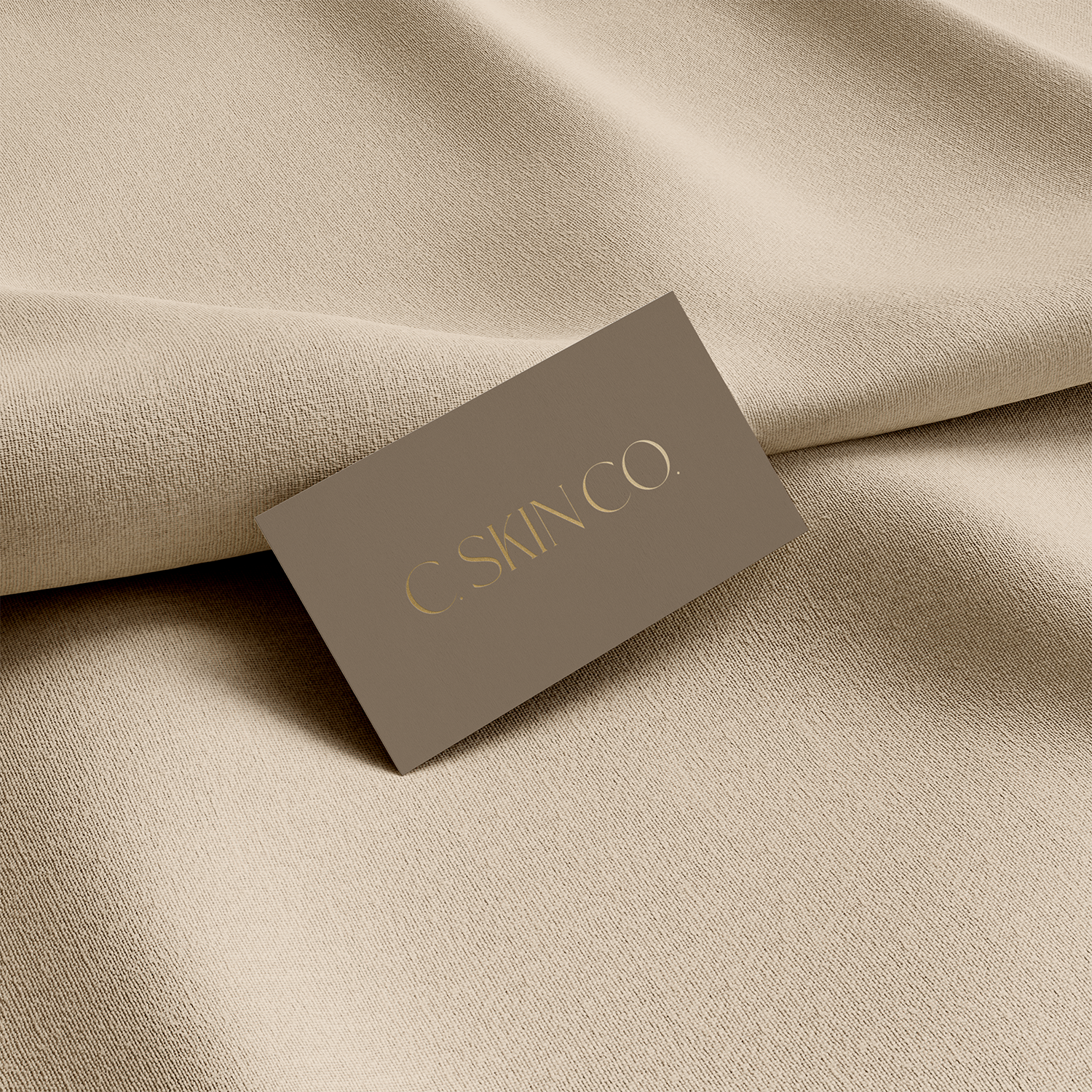 A beige fabric surface with a card reading "C. SKIN CO." placed on top, showing a minimalistic skincare brand presentation.