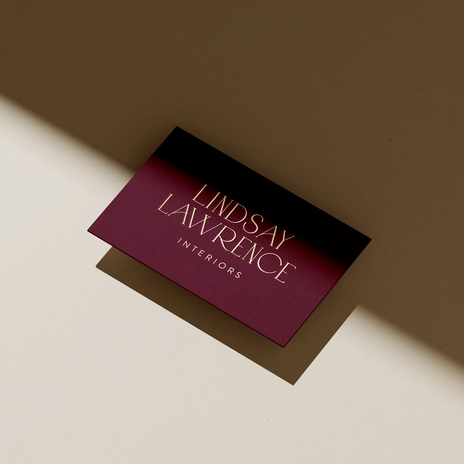 A business card for Lindsay Lawrence Interiors with a maroon and black gradient background and white text, placed on a beige surface with its shadow cast to the left.