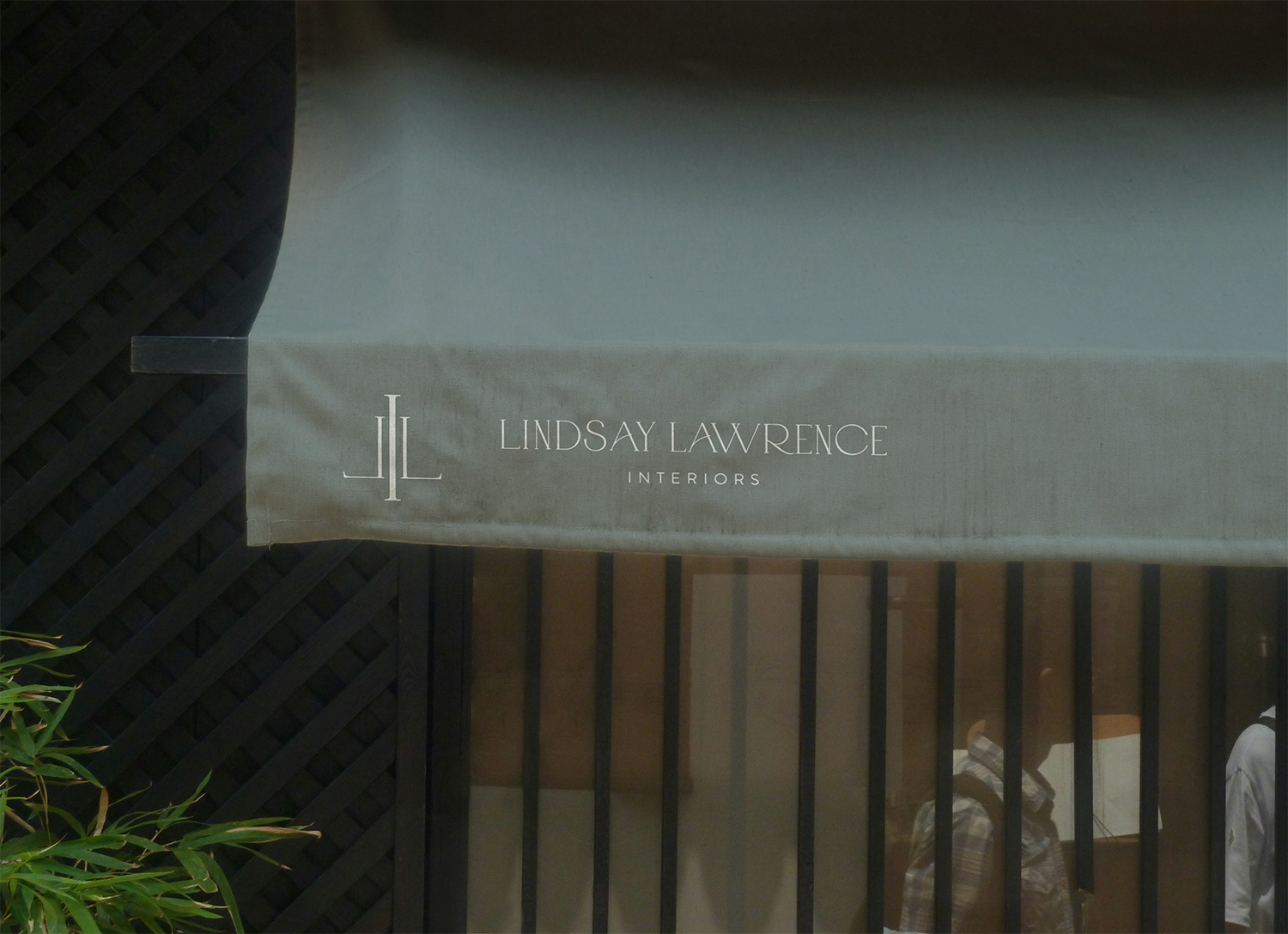 Sign reading "Lindsay Lawrence Interiors" on a beige fabric banner outside a building with windows and black fence.