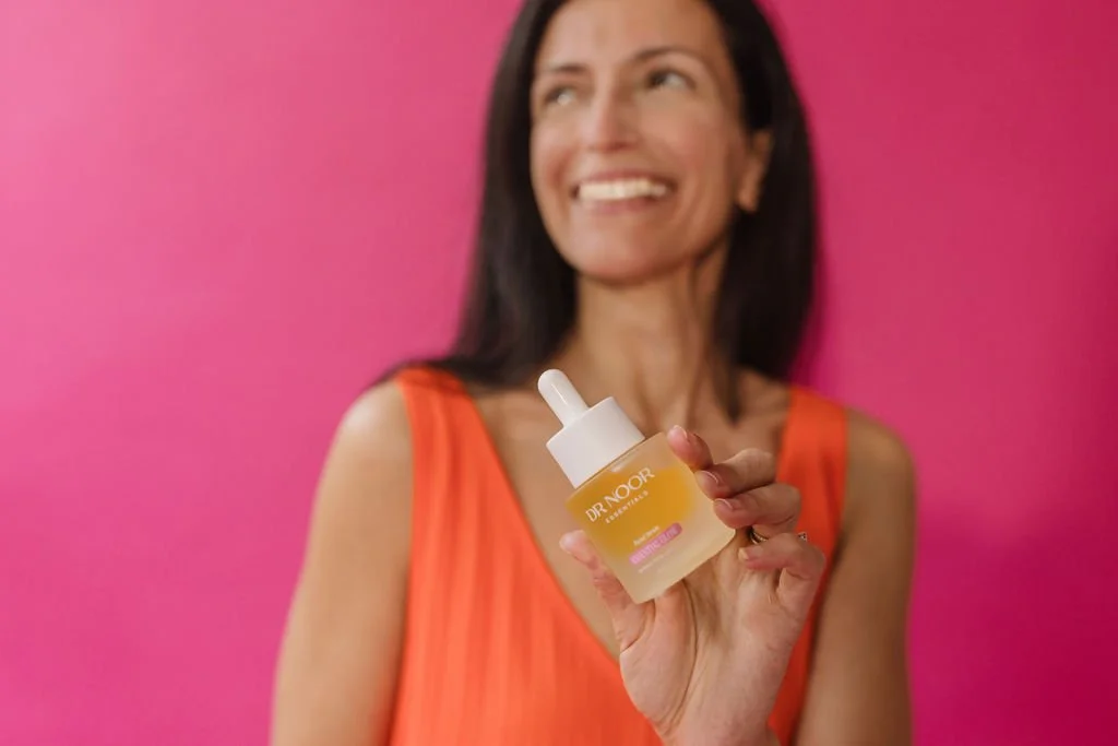 Woman with dark hair smiling, holding a skincare serum bottle labeled 'Dr Nokor' against a pink background.
