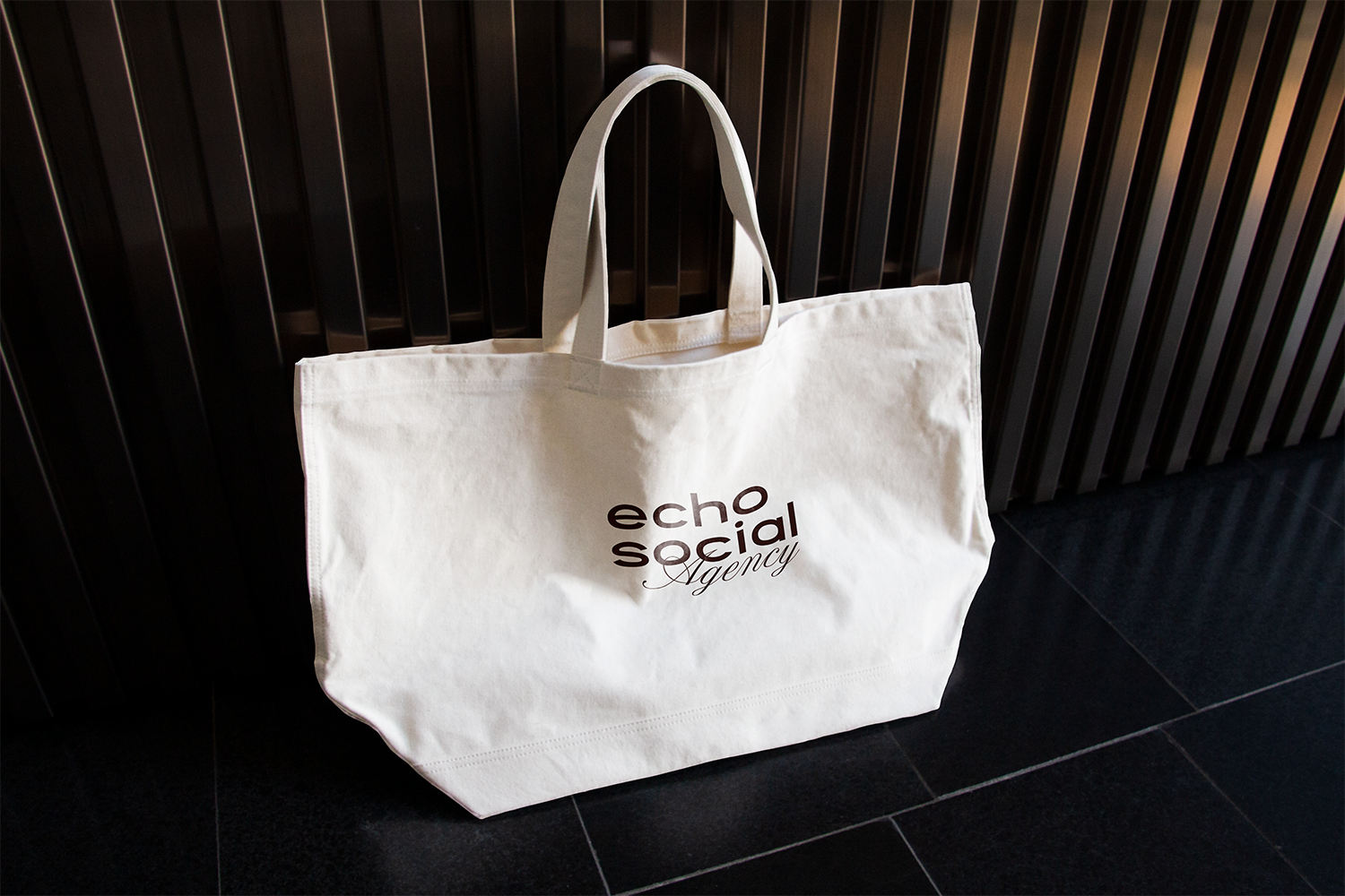 A white tote bag with the words 'echo social Agency' printed on it, leaning against a dark wall and resting on a tiled floor.
