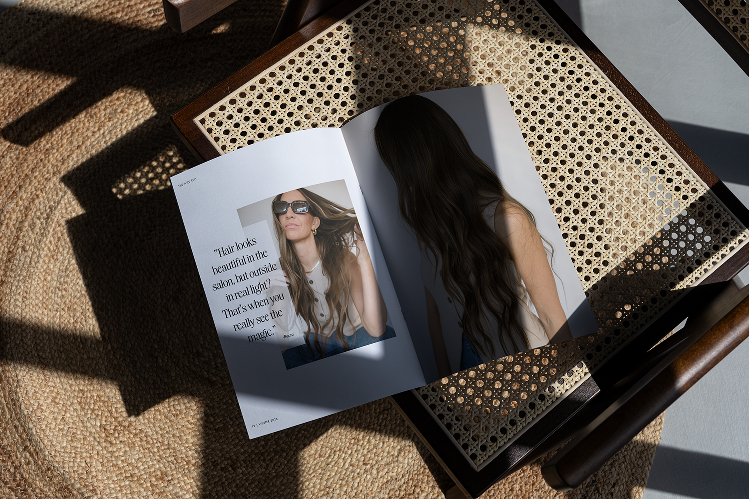 Open magazine on a wooden and woven rattan table showing a woman with long wavy brown hair wearing sunglasses, and another photo of her with face hidden by hair, indoor sunlight casting shadows.