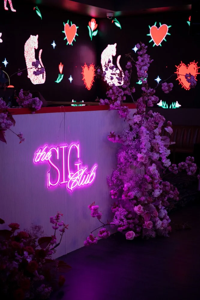 Neon pink sign reading 'the SIG Club' with pink flowers and a floral arrangement in front, and a dark background with animated hearts and cats on a screen.