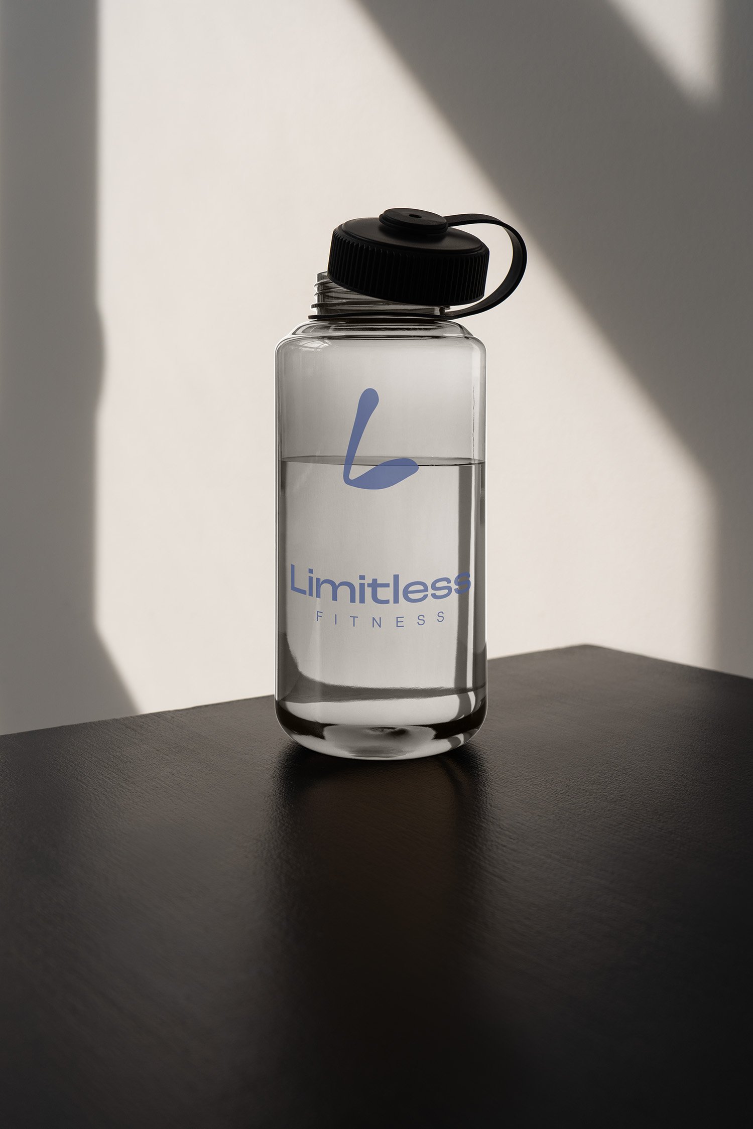 A clear water bottle with a black lid and a handle, featuring the words 'Limitless Fitness' in blue, placed on a dark surface with shadows and sunlight in the background.