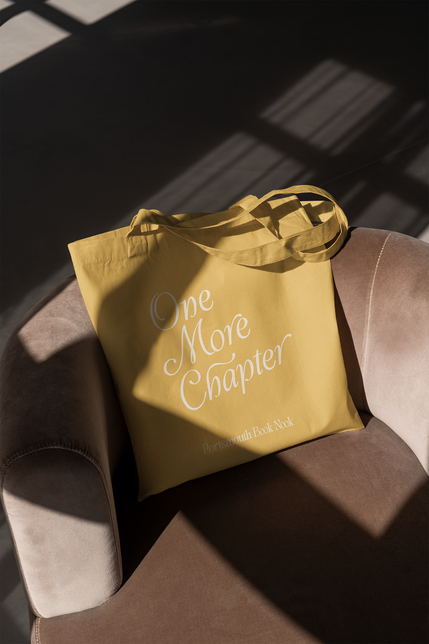 Yellow tote bag with white text resting on a beige armchair, with shadows cast by a window on the background.