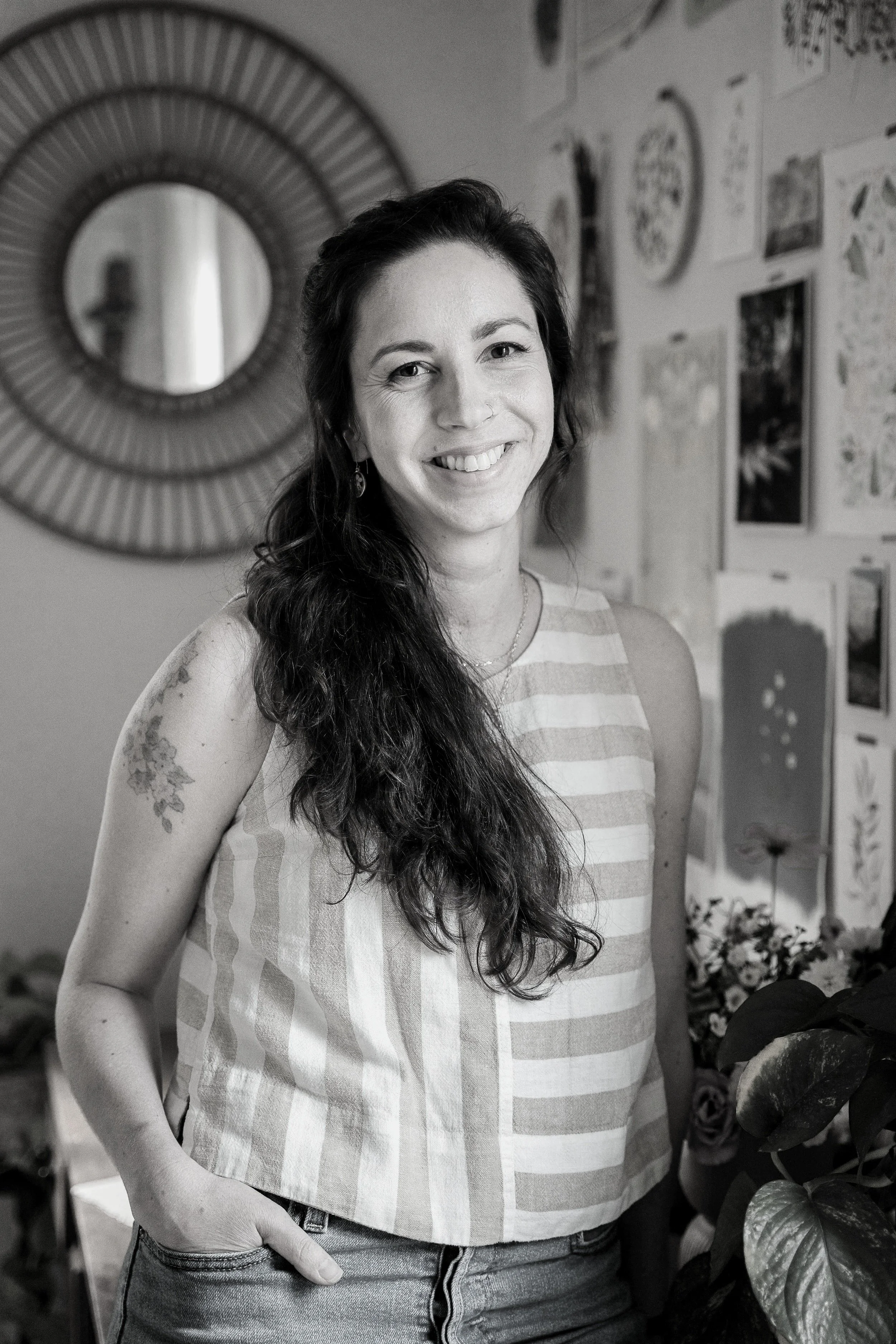 Black-and-white photo of a smiling woman with long, wavy hair wearing a sleeveless striped top and jeans, standing indoors near a wall with decorative artwork and a round mirror.