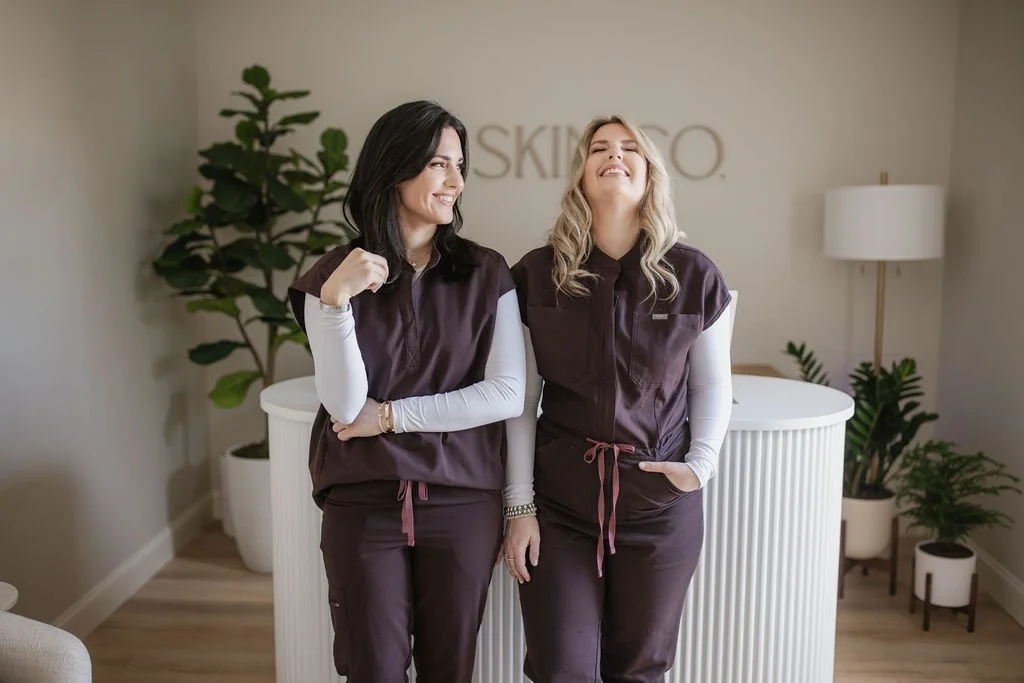 Two women in matching spa uniforms smiling and talking in a modern spa reception area with plants and a white counter.