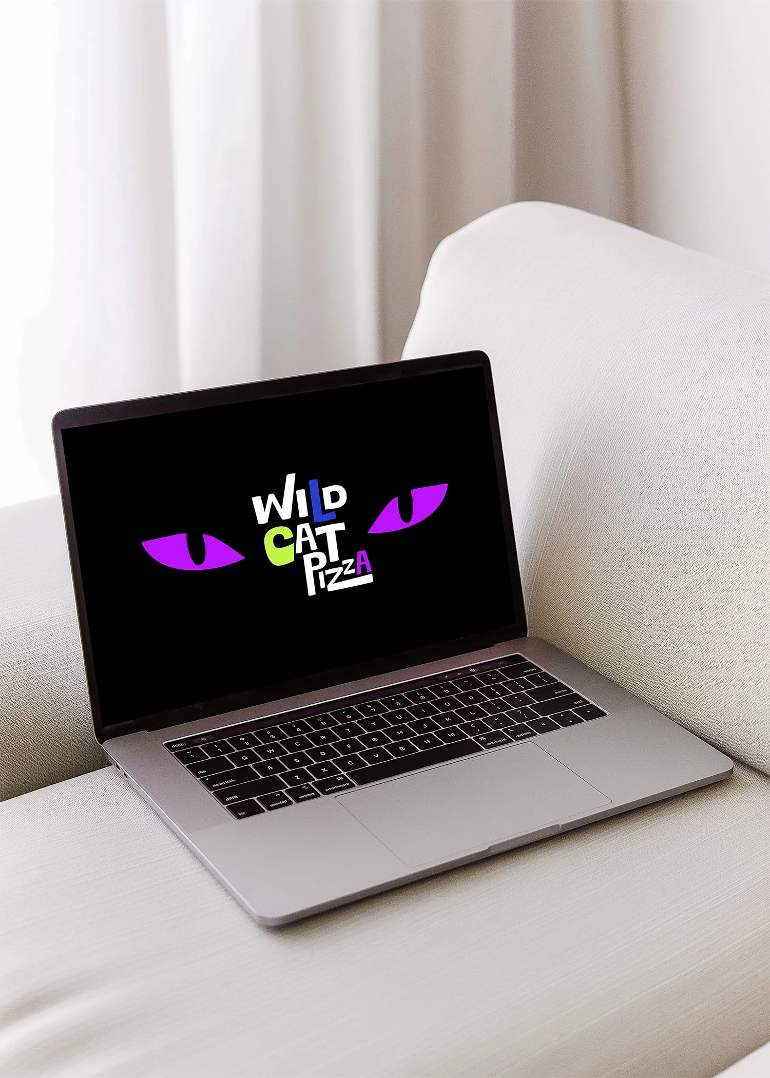 A silver MacBook laptop on a white table, displaying the logo for Wild Cat Pizza, a black background with purple cat eyes and the words 'Wild Cat Pizza' in colorful letters.